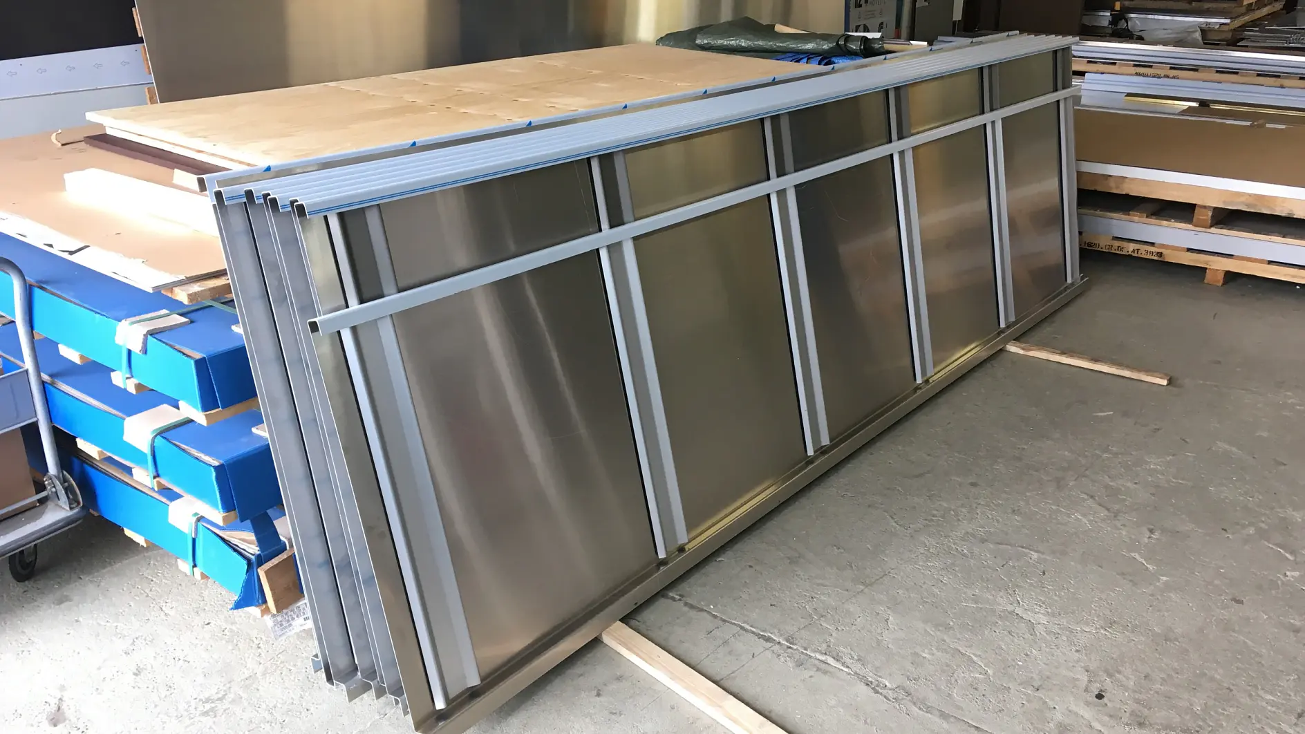 Stacks of large metal panels are leaning against a table in a warehouse. Nearby, there are boxes on a cart and other materials, including rolls of tesa tape. The scene is well-lit, with a concrete floor and industrial surroundings. (This text has been generated by AI)
