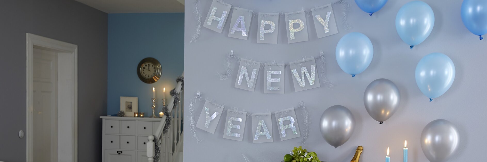 A room decorated for New Years Eve with a Happy New Year banner on the wall, secured using tesa tape. Blue and silver balloons are displayed. A table holds a champagne bucket, glasses, candles, and a bowl of fruits. A white cabinet and staircase are visible in the background.