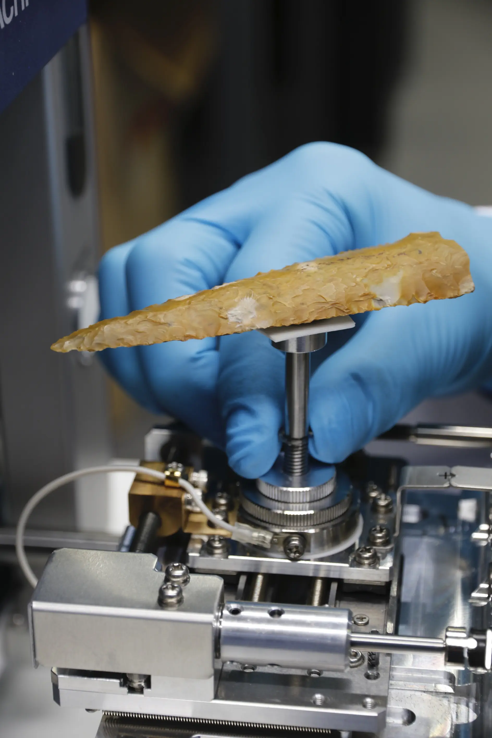 A gloved hand holding a triangular, serrated stone tool above a metallic apparatus, likely designed for scientific analysis. The stone appears to be an ancient or archaeological artifact secured in place using tesa tape.
