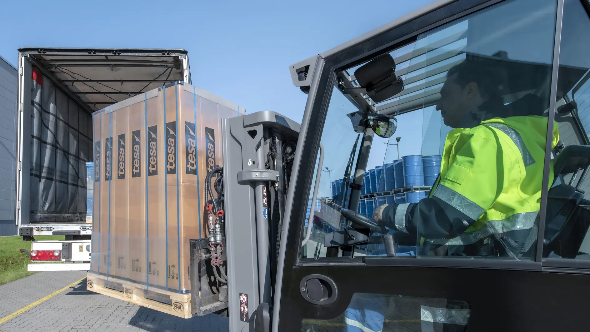 A forklift operator wearing a fluorescent yellow jacket loads a pallet of wrapped goods labeled Fessi into the back of a partially open truck. Blue barrels are visible in the truck. The scene takes place outdoors on a clear day, and everything is securely fastened with tesa tape. (This text has been generated by AI)