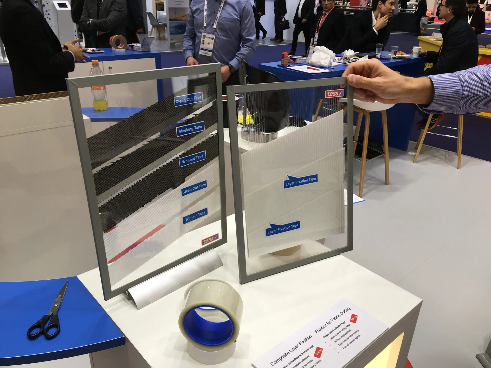 A person holds a mesh screen with labeled samples of various tesa tapes. The table displays a roll of blue tesa tape, scissors, and product information. People converse in the background at a trade show or similar event.