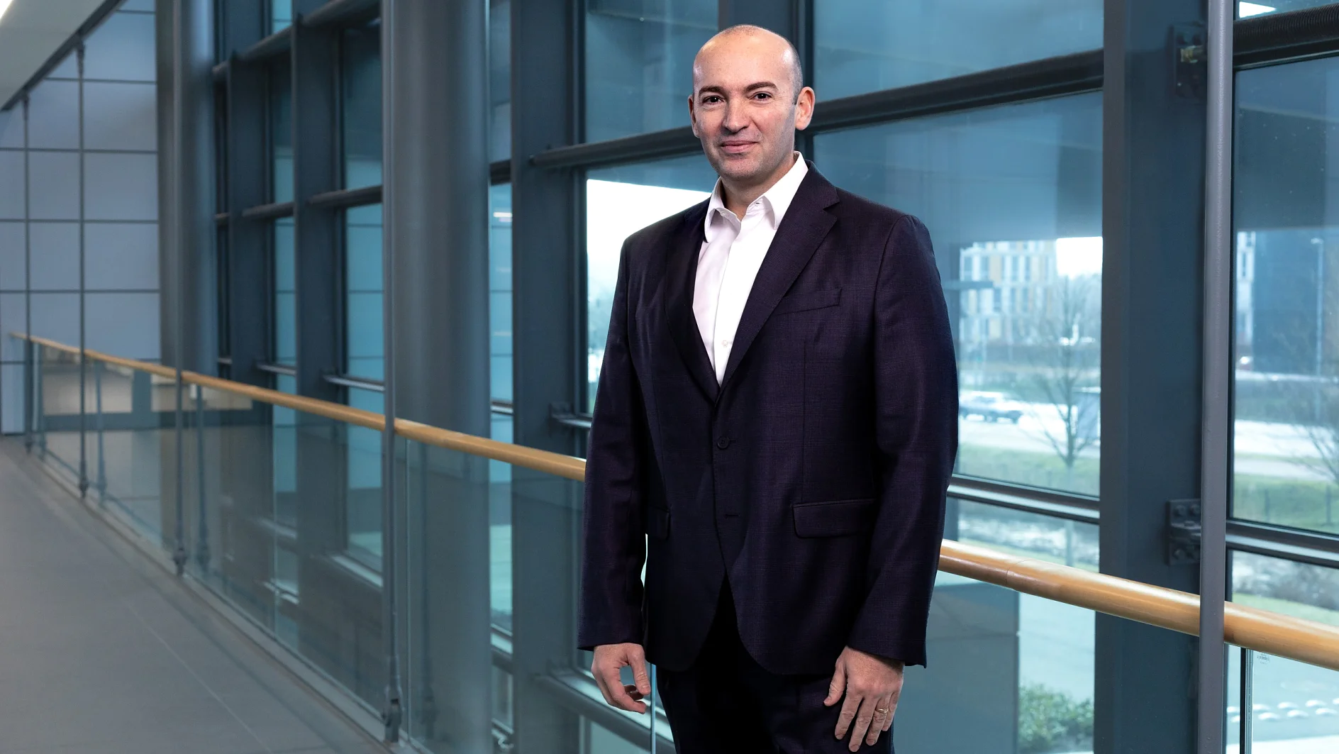 A man in a dark suit stands in a modern hallway with large glass windows, allowing natural light to stream in. The landscape outside is partially visible, showing buildings and greenery. (This text has been generated by AI)