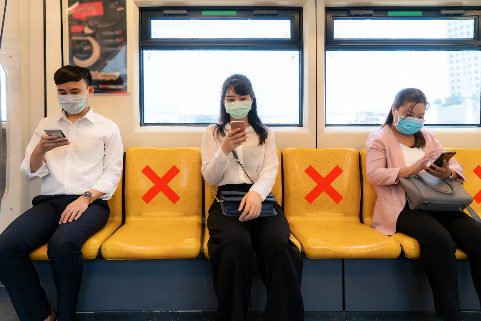 Three people wearing masks sit on a train with yellow seats. Red X marks, made with tesa tape, indicate seats meant for social distancing. Each person is using their phone. Two windows are visible in the background. (This text has been generated by AI)