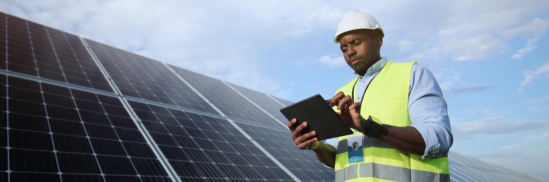 A person wearing a hard hat and neon safety vest is using a tablet near large solar panels secured with tesa tape. The sky is partly cloudy.
