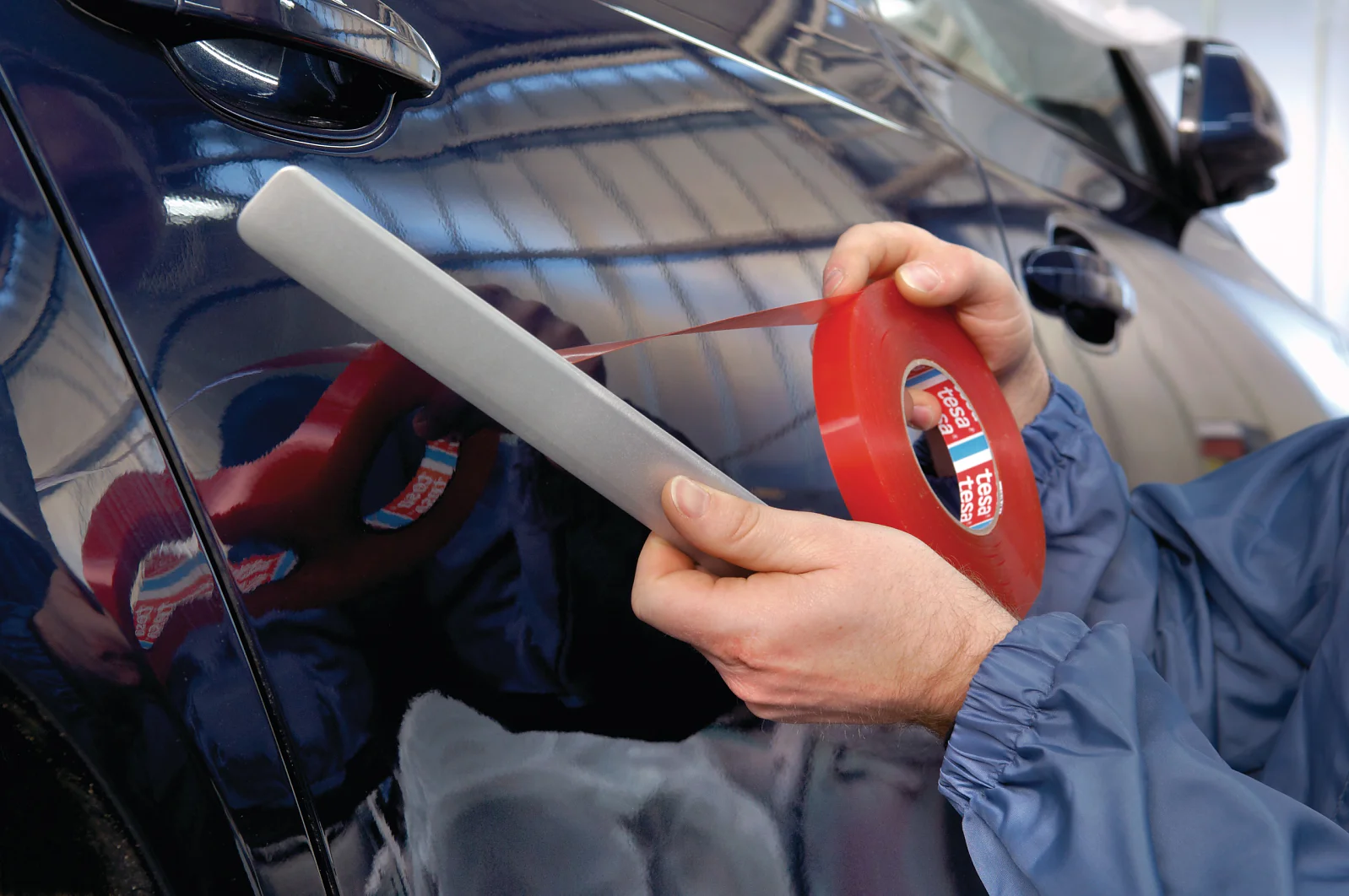 A person in a blue jacket is applying red tesa tape to the side of a dark-colored car. They are holding a tool with the tesa tape to ensure even application. The background shows a blurry interior of a garage or workshop. (This text has been generated by AI)