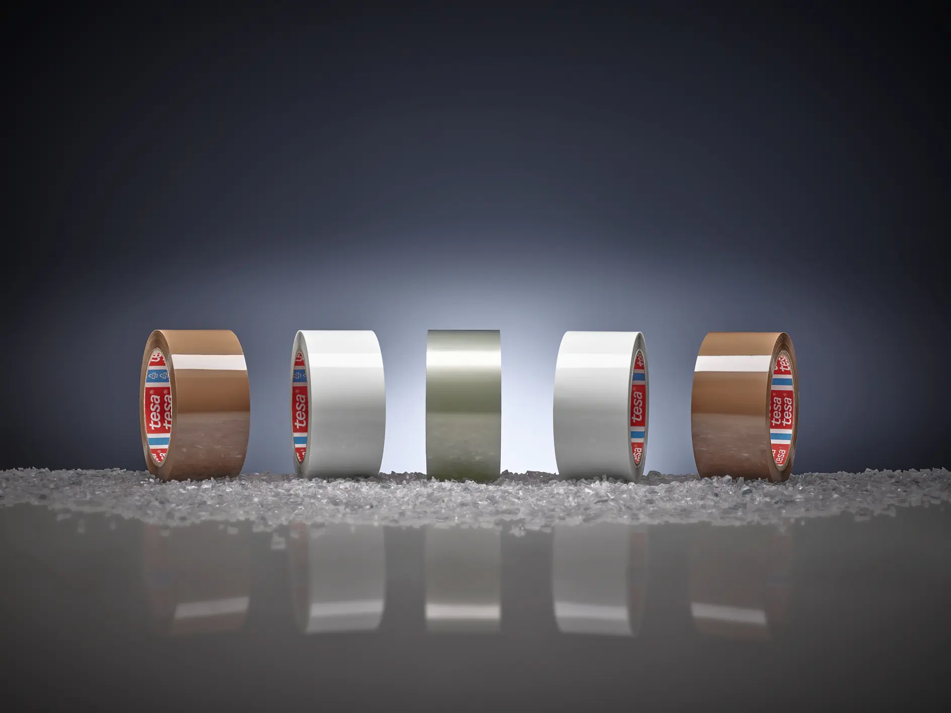 Four rolls of adhesive tesa tape, two brown and two white, are lined up on a surface scattered with small granules. Each roll is standing upright in front of a dark gradient background. (This text has been generated by AI)