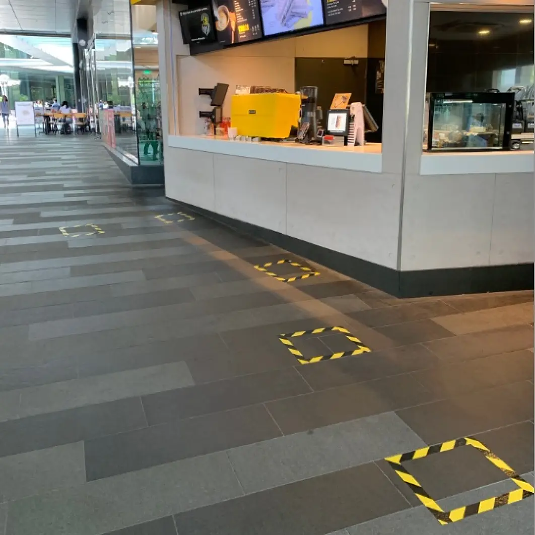 A cafe counter with a digital display menu is on the right. Yellow and black tesa tape marks distancing squares on the gray tiled floor. Seating is visible in the background to the left. (This text has been generated by AI)