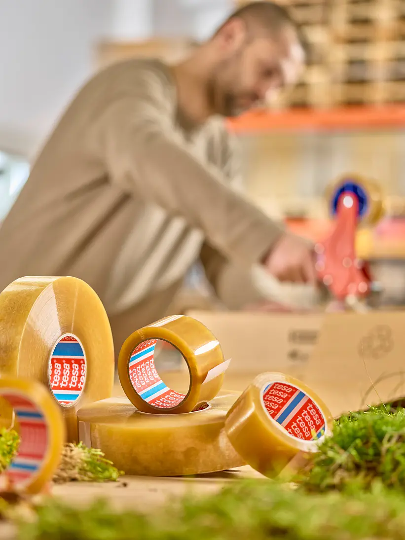 A person in a beige sweater is using a tesa tape dispenser in a warehouse setting. Several rolls of tesa tape are in the foreground, surrounded by patches of greenery. Shelves with boxes and pallets are visible in the background. (This text has been generated by AI)