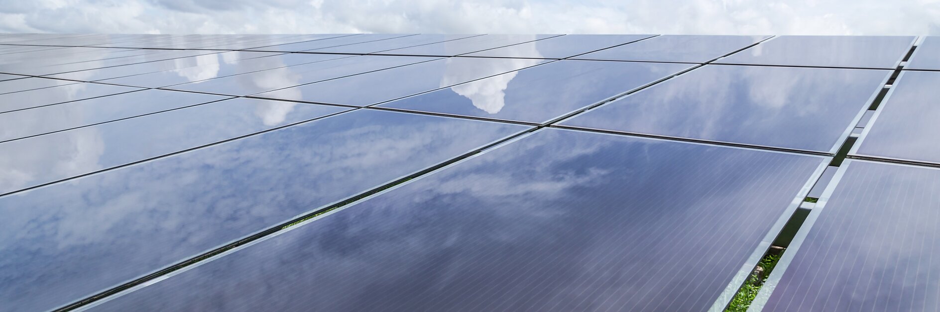 A large array of solar panels is seen reflecting a partly cloudy sky. The panels stretch across the image, capturing a mix of blue sky and white clouds. Vegetation is visible between the panels.