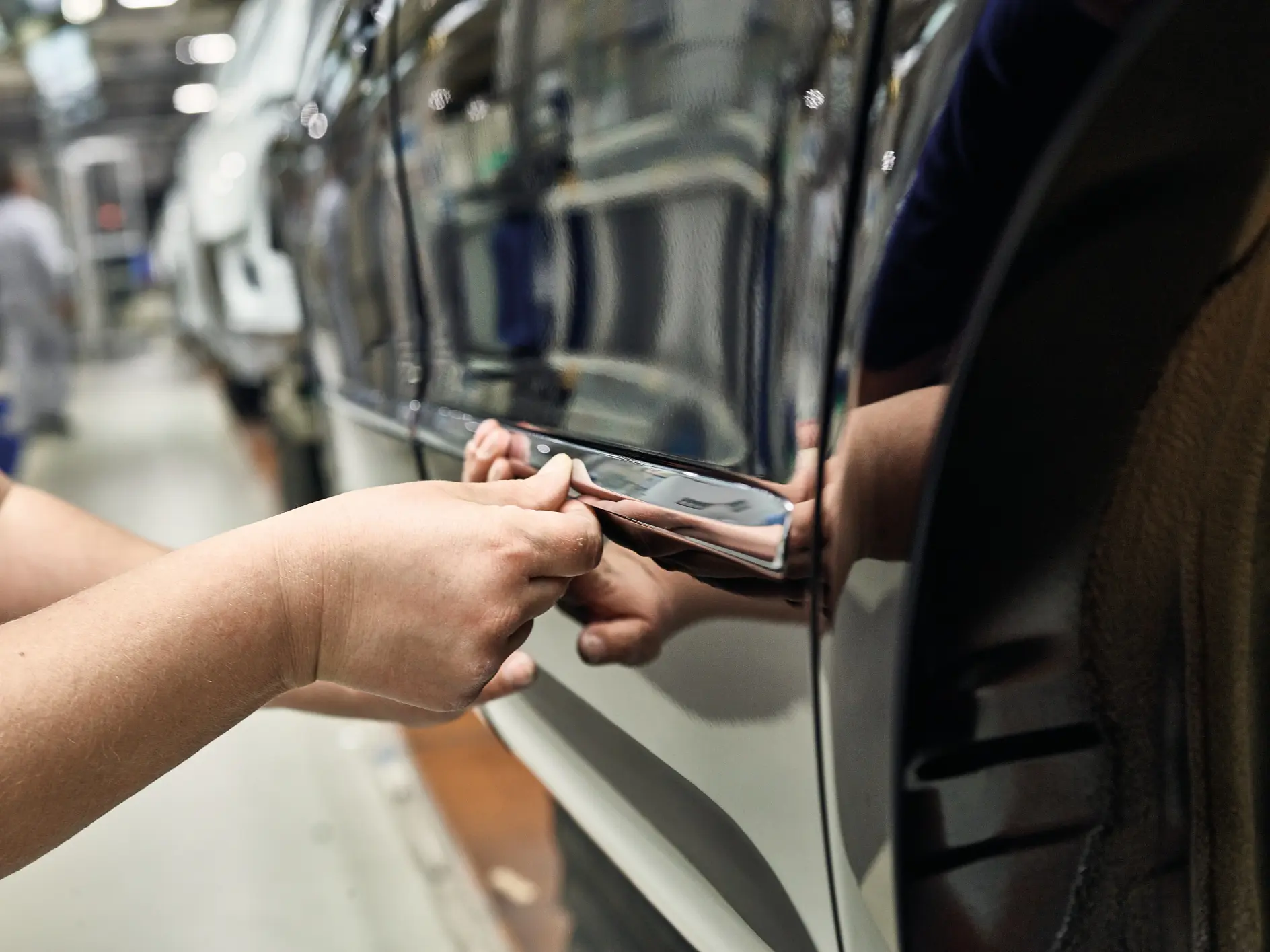 Close-up of a person’s hands applying a chrome strip to the side of a car on a production line using tesa tape. The cars glossy surface reflects the surrounding factory environment. Another person is visible in the background, out of focus.
