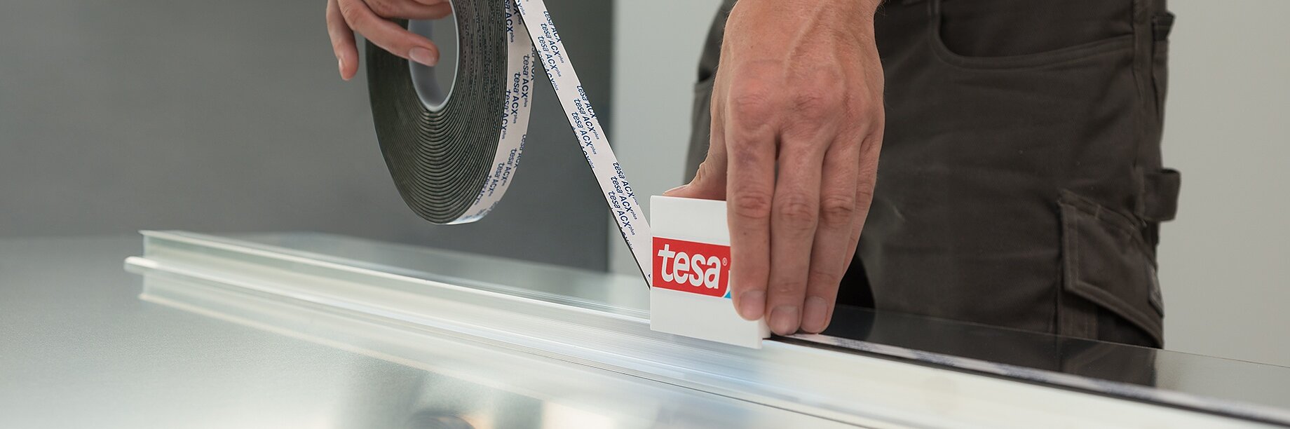 A person applies tesa tape to a metallic surface. The tesa tape is being pressed onto the surface using a plastic tool. The person is wearing a black shirt and khaki pants.