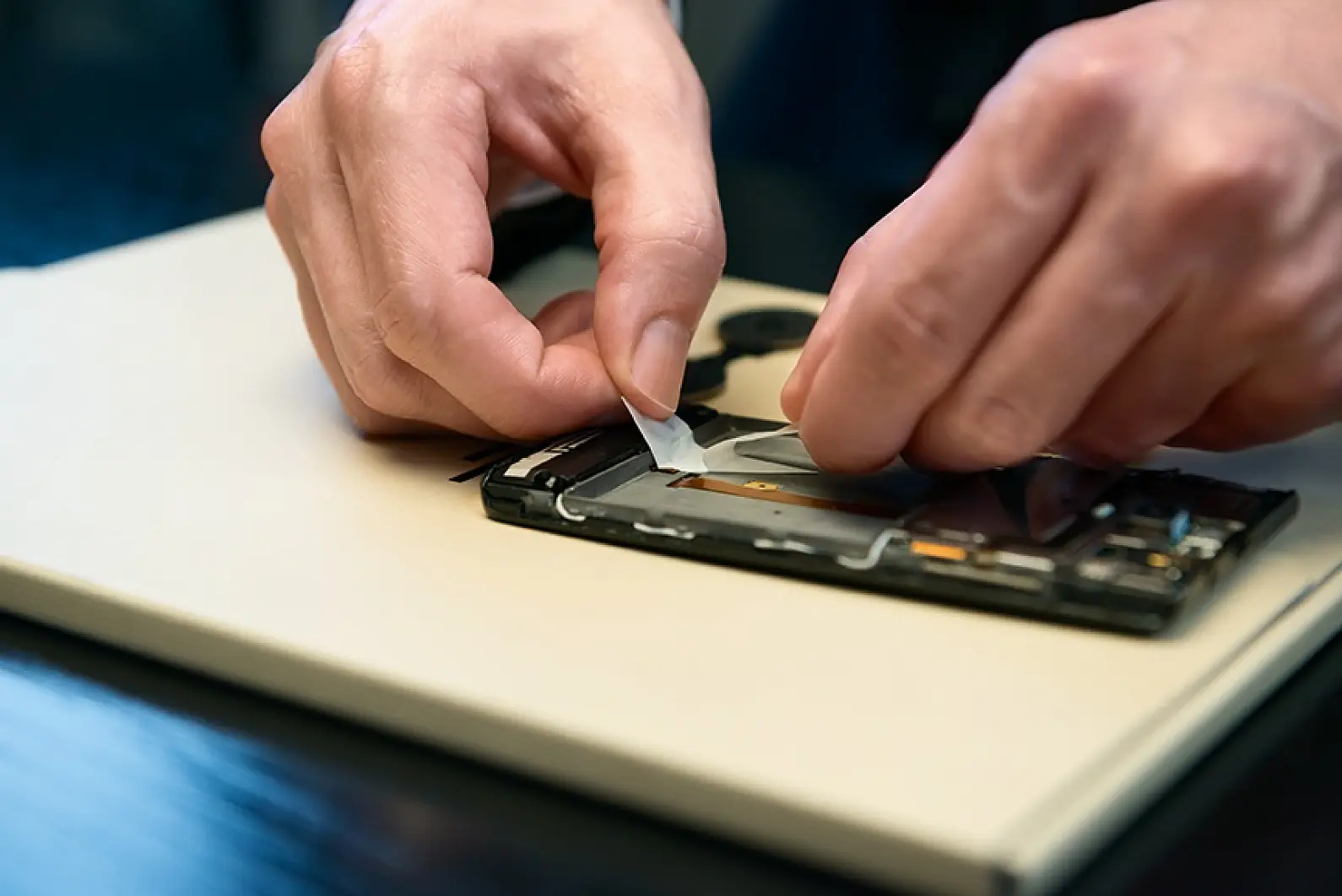 Two hands carefully work on disassembling a smartphone on a table. The person is removing a component from the device, which has its back cover open, revealing internal parts. The background is blurred, focusing on the hands and the phone secured with tesa tape. (This text has been generated by AI)