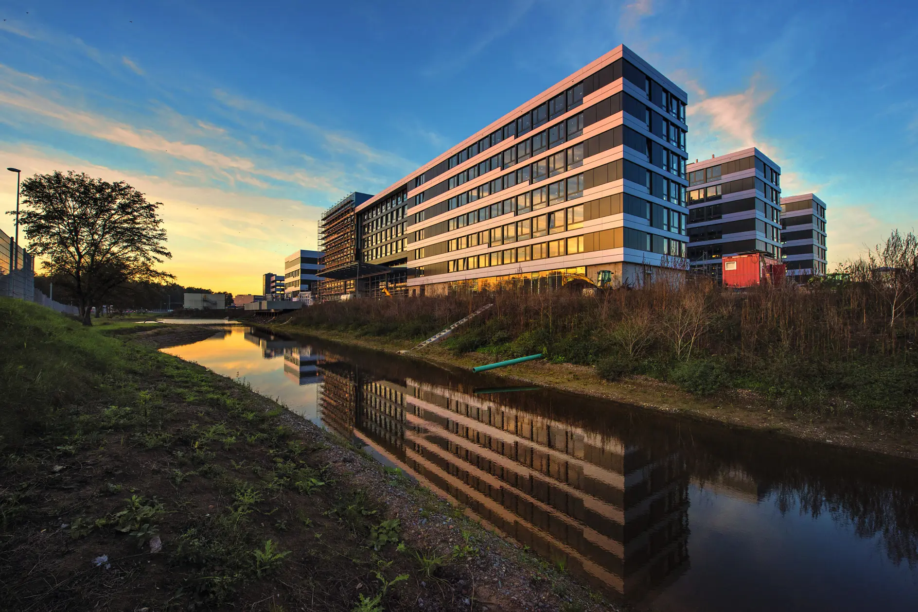 Modern glass and concrete buildings are reflected in a calm canal during sunset. The sky is painted with shades of blue, orange, and yellow. A grassy area lines the water, and trees are visible in the background.