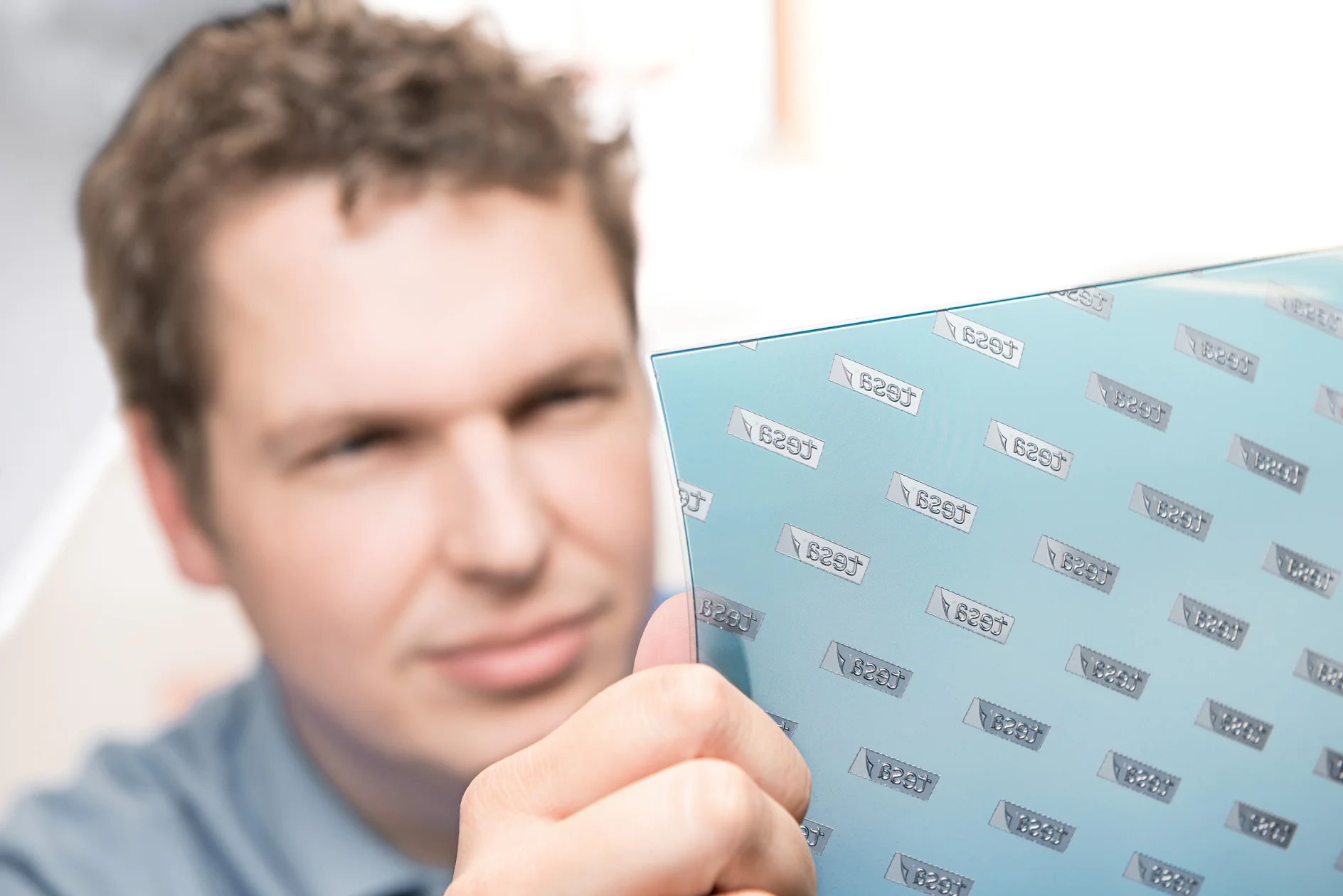 A man holds a transparent, reflective sheet at an angle. The sheet is covered with the word tesa tape repeated in a pattern. The mans face is slightly out of focus, while the sheet is in focus, reflecting light. (This text has been generated by AI)