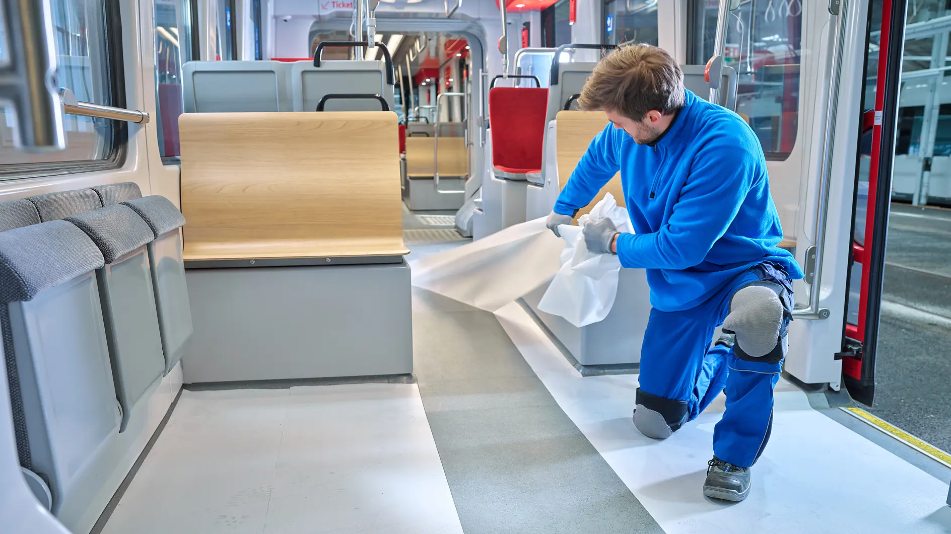 A person in a blue uniform kneels on the floor of a train, applying white tesa tape sheets. The train interior features gray flooring, wooden benches, and red seats. The person is wearing knee pads and gloves.