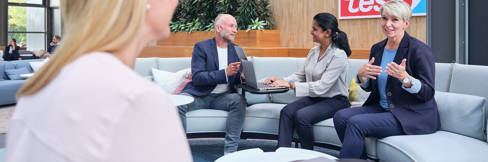 Four people sit in a modern office lounge, engaged in conversation. Laptops and documents are on the table. A tesa logo is on the wall. A digital display in the background shows an image and text. (This text has been generated by AI)