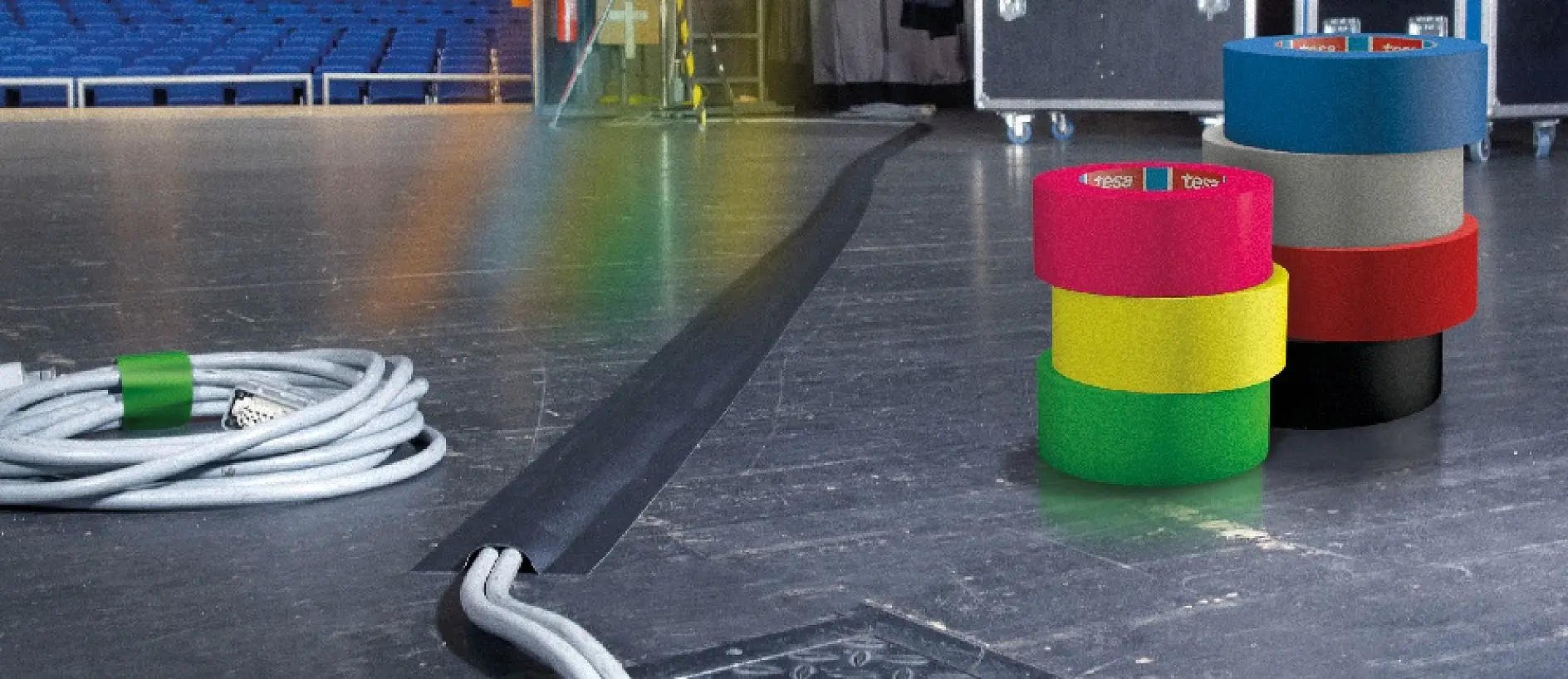 A stage with black tesa tape securing a cable to the floor. Nearby, several rolls of colored tesa tape in pink, red, green, yellow, blue, gray, and black are stacked. Coiled cables with a green band are on the left.