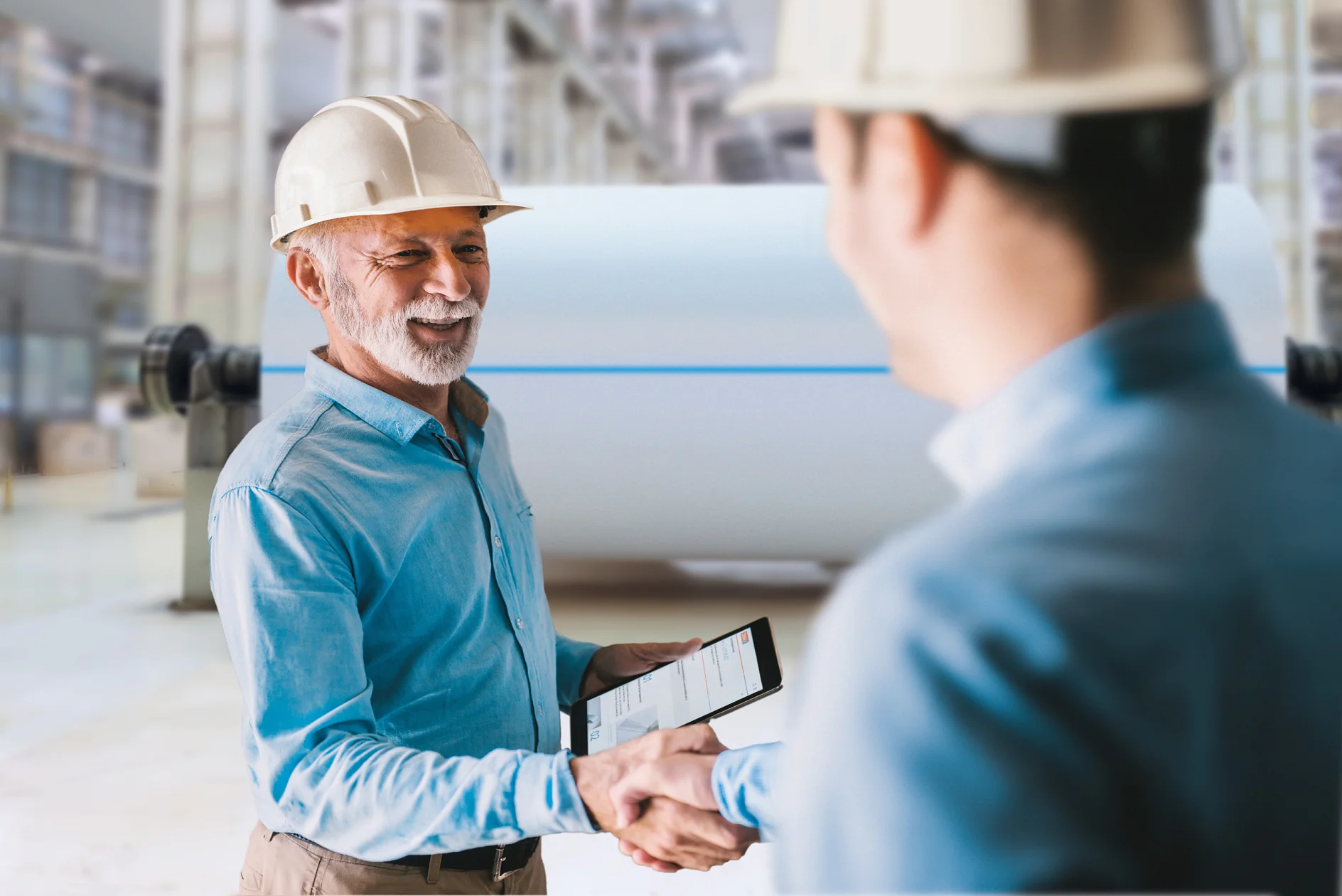 Two men wearing safety helmets shake hands in an industrial setting. One holds a tablet displaying graphs. Large machinery is blurred in the background, suggesting a factory or manufacturing environment, where tesa tape might be commonly used for various applications. (This text has been generated by AI)