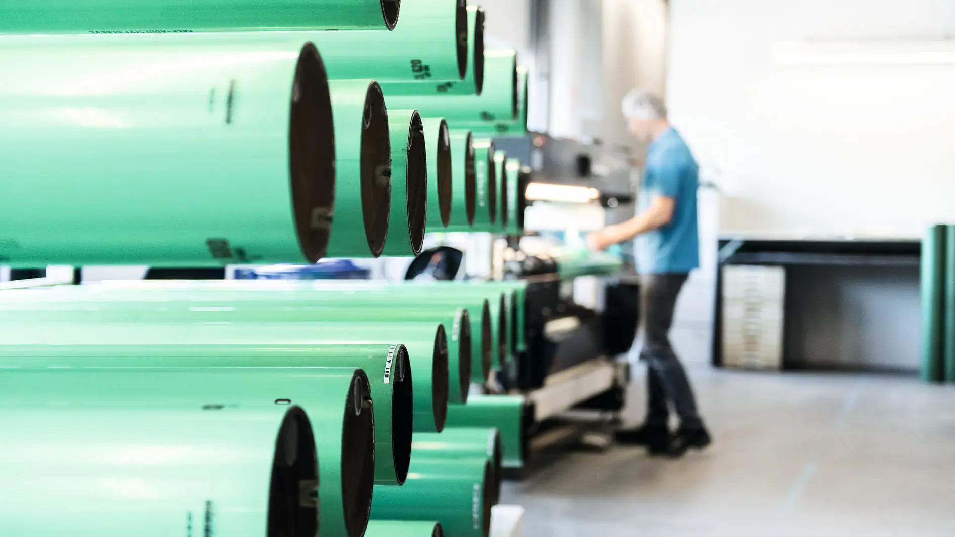 Rows of large green cylindrical tubes are lined up in a workshop. A person wearing a blue shirt is blurred in the background, working with tesa tape and other equipment. The setting appears to be an industrial or manufacturing environment.