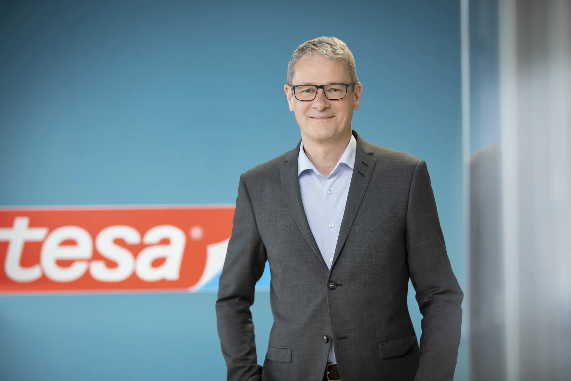 A man with gray hair and glasses, dressed in a gray suit and light blue shirt, stands in front of a blue wall with a red and white tesa tape sign partially visible.