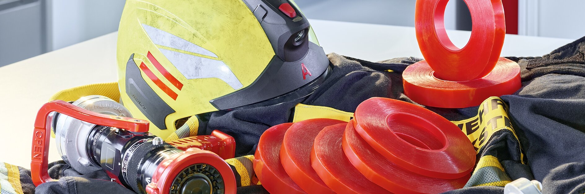 A firefighters helmet and hose are displayed with several red sealing rings on a yellow and black fabric. In the background, a fire extinguisher is visible through an open door.