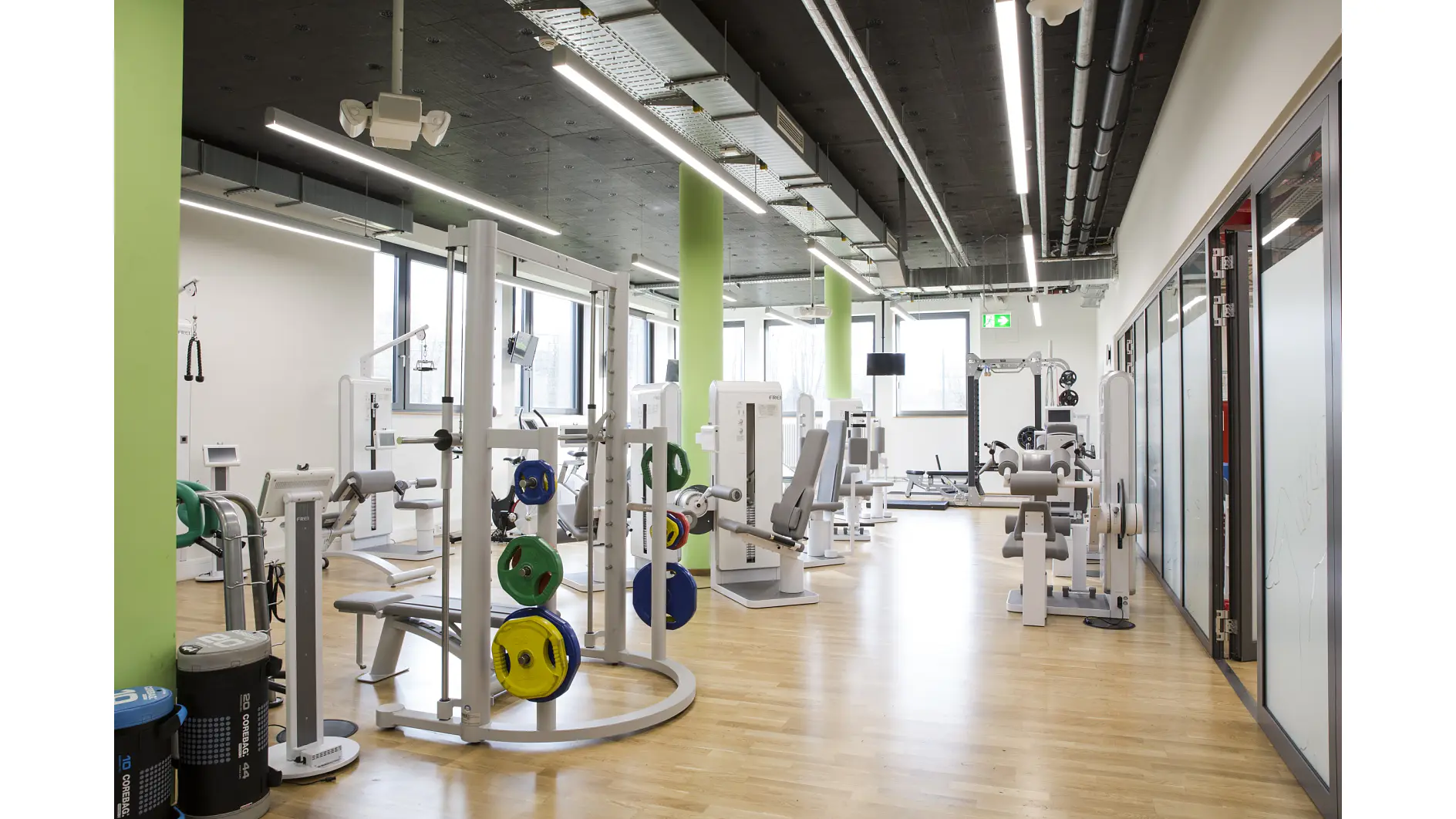 A modern gym with wooden floors and green accents features various exercise equipment, including weight machines and free weights secured using tesa tape. There are large windows on one side, and the space is well-lit with overhead lights. (This text has been generated by AI)
