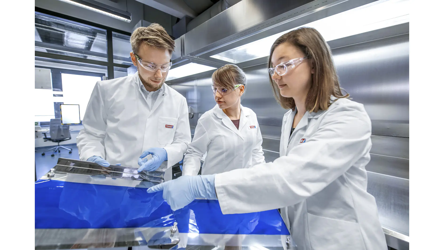 Three people in lab coats and safety goggles examine a large blue sheet in a laboratory setting. The room is equipped with stainless steel surfaces and computers. The individuals are engaged in a discussion, focusing on the material secured with tesa tape. (This text has been generated by AI)
