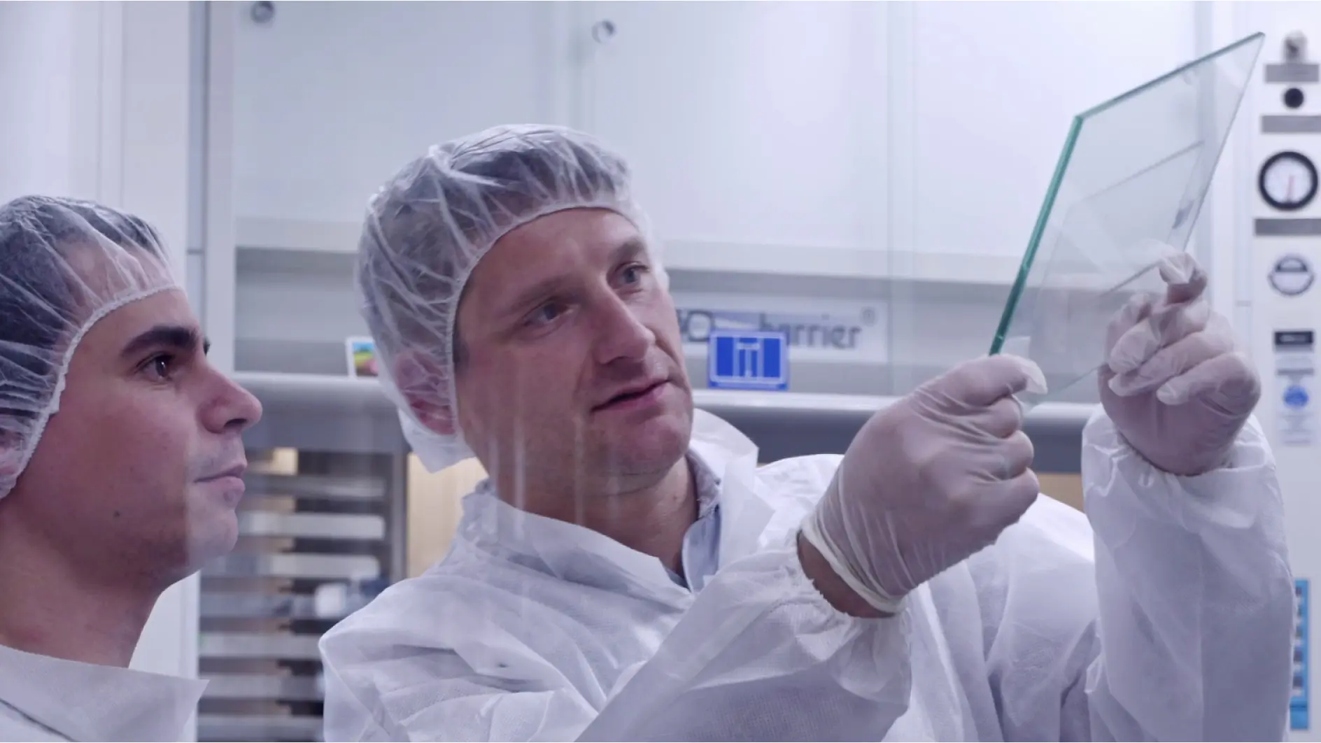 Two people in protective lab clothing and hairnets, one holding and examining a transparent sheet of tesa tape. They appear to be in a laboratory setting with equipment visible in the background. (This text has been generated by AI)