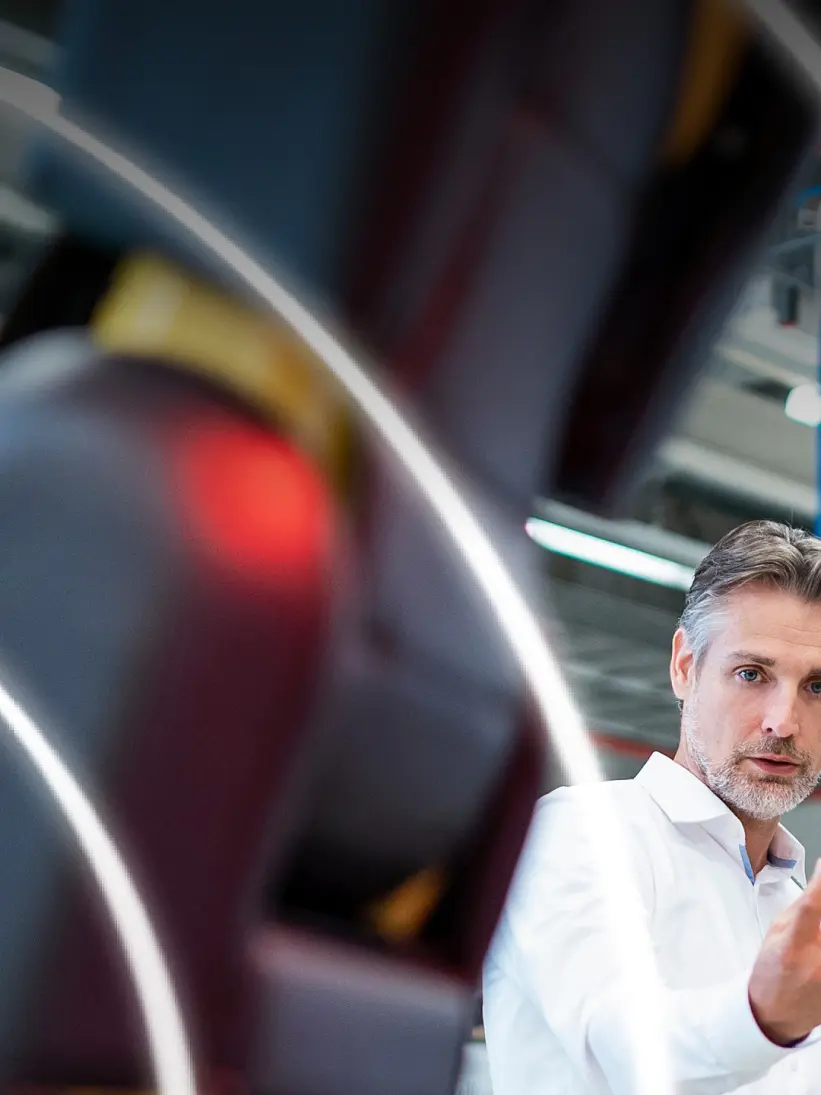 Two people in a modern industrial setting observe a large robotic arm in the foreground. The man gestures with his hand, while the woman stands beside him, both appearing engaged with the machinery. Visible are overhead cables and lighting secured with tesa tape. (This text has been generated by AI)