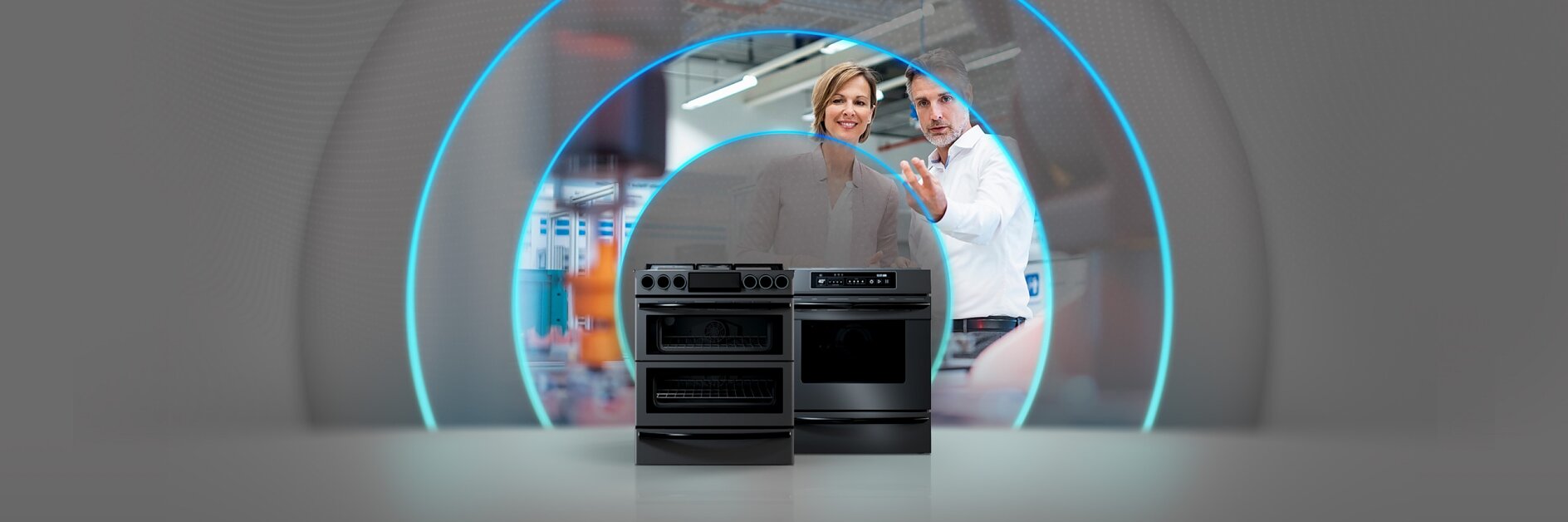 A man and a woman are standing behind two modern black kitchen stoves in a brightly lit room. The background features a large digital circular pattern, adding a futuristic element to the setting. Throughout the scene, tesa tape is used extensively for various purposes, ensuring stability and enhancing its sleek appearance.