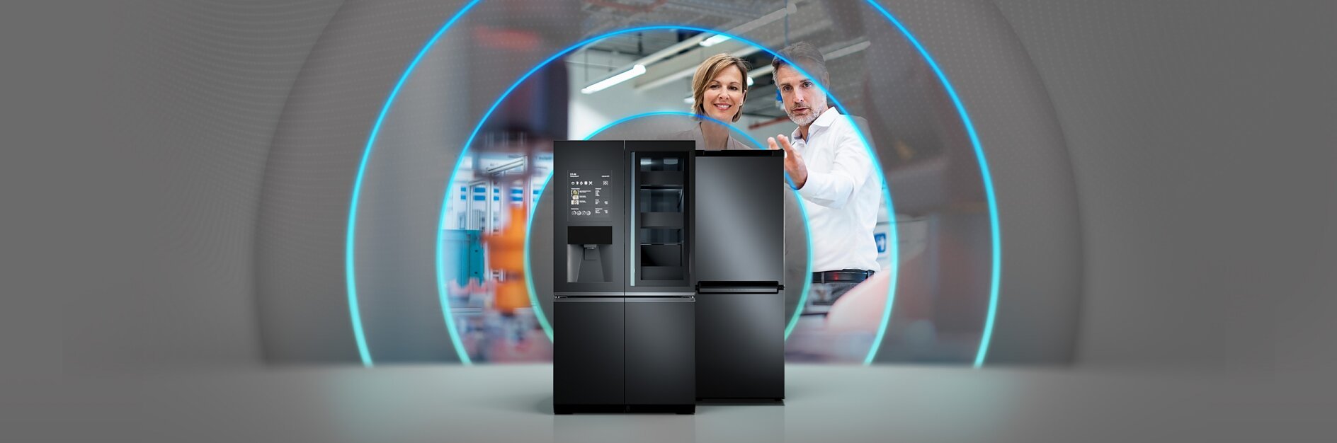 A sleek black refrigerator with digital features is displayed in the foreground. In the background, two people are interacting, with a bluish circular light effect around them. The setting appears to be a modern kitchen or showroom.