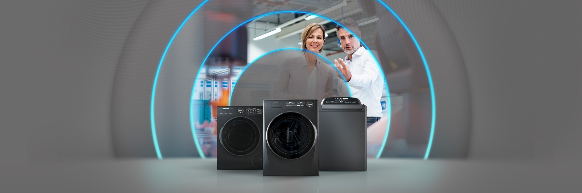 Three black home appliances are displayed in the foreground, possibly washing machines or dryers. In the background, two people wearing business attire appear to be discussing or presenting the appliances, with a digital overlay of blue arcs surrounding them.