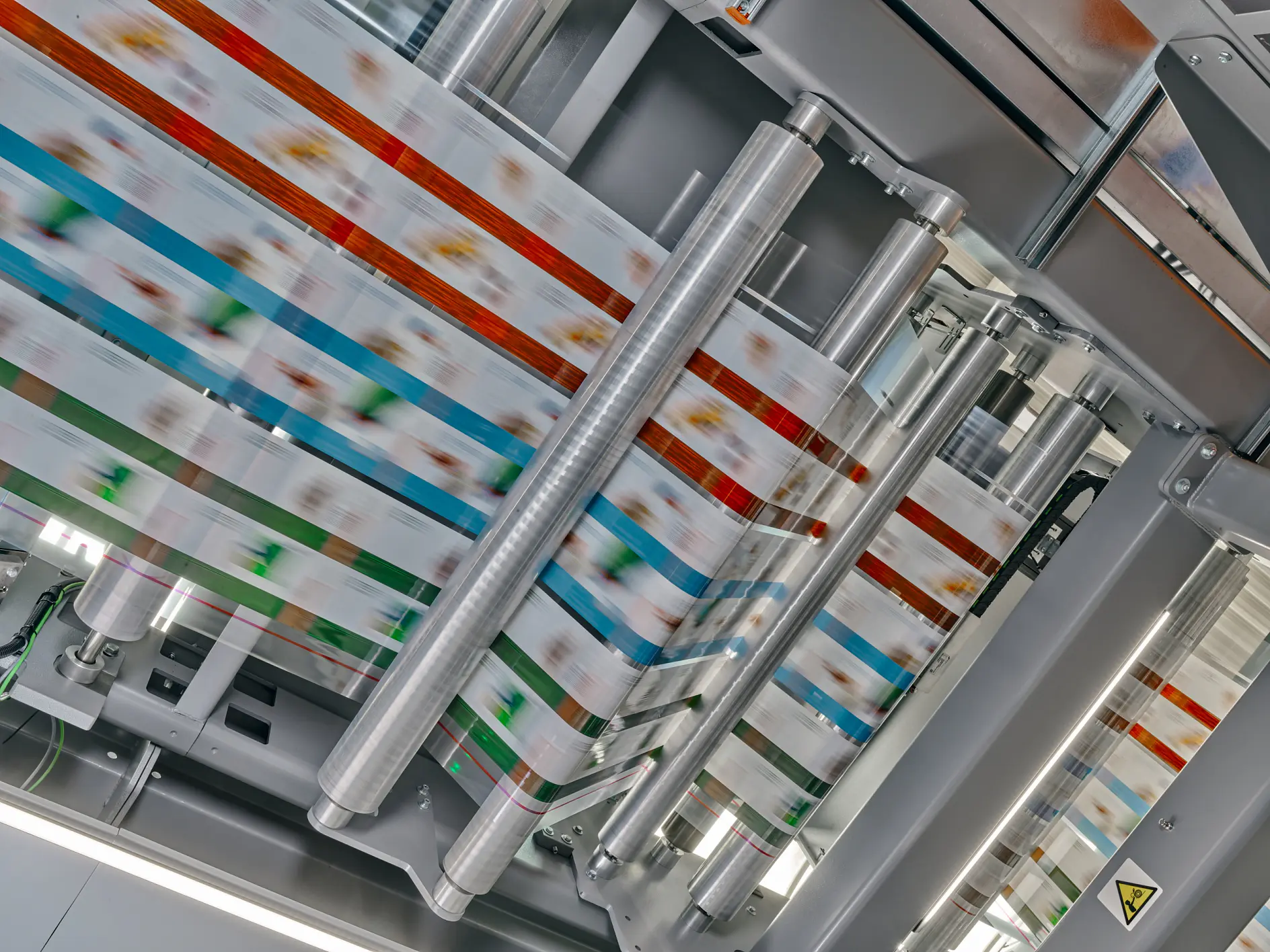 A high-speed printing press produces colorful packaging material. Multiple rollers guide the printed material through the machine, showing a blur of colors, including red, blue, and green patterns, under bright lighting. The process relies on the use of tesa tape to ensure everything stays in place during production. (This text has been generated by AI)
