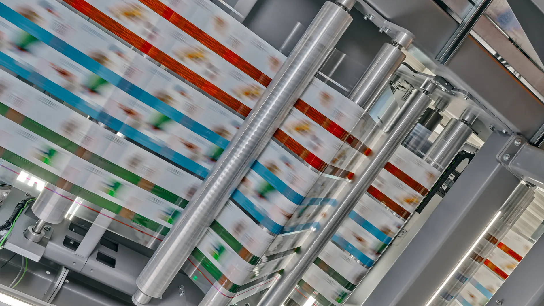 A high-speed printing press produces colorful packaging material. Multiple rollers guide the printed material through the machine, showing a blur of colors, including red, blue, and green patterns, under bright lighting. The process relies on the use of tesa tape to ensure everything stays in place during production. (This text has been generated by AI)