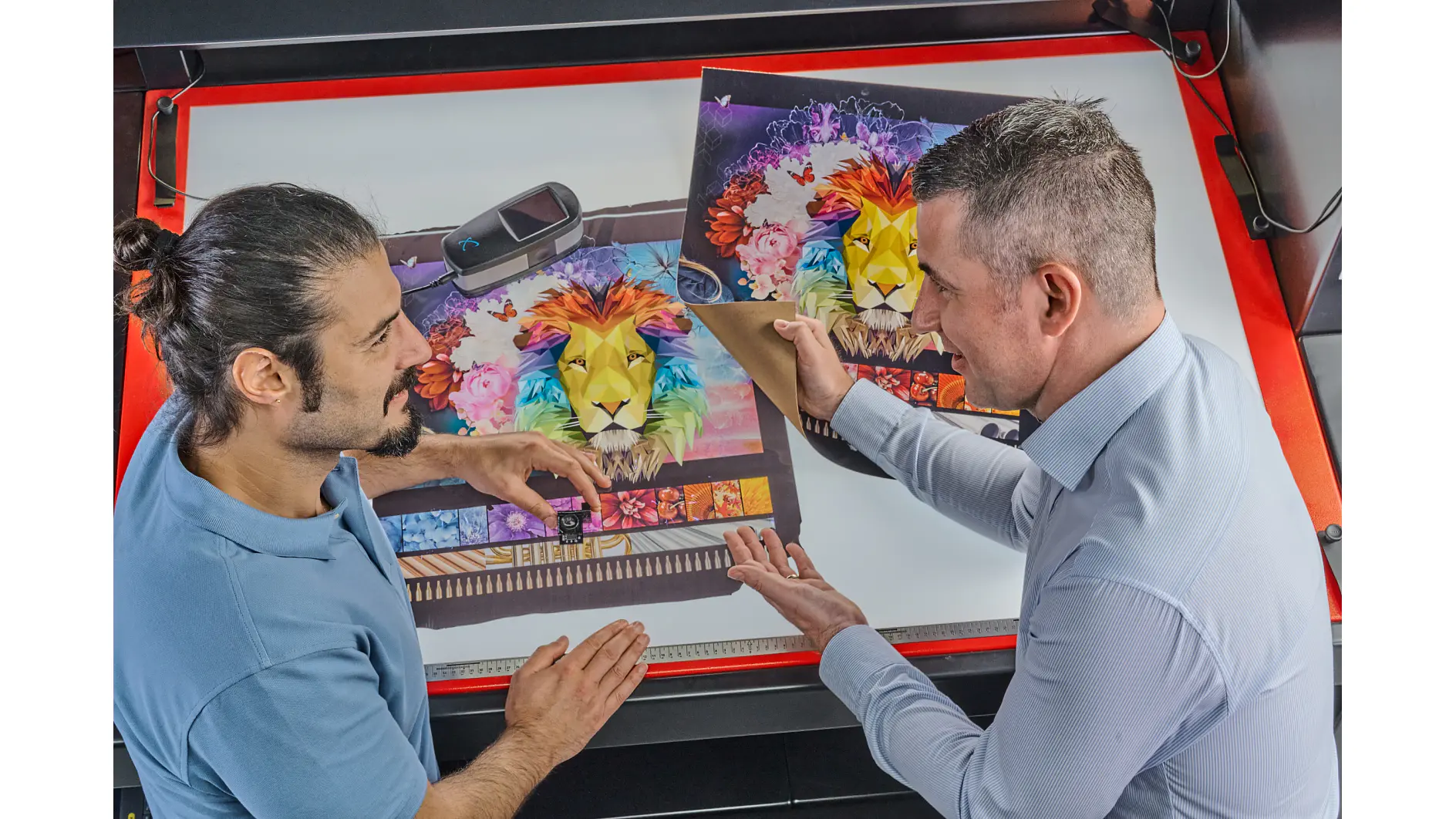 Two men are discussing a vibrant print of a lion surrounded by colorful flowers. One man holds the print, while the other gestures towards it. A color calibration device is on the table beside them, along with some tesa tape used to secure edges and ensure everything stays in place. (This text has been generated by AI)