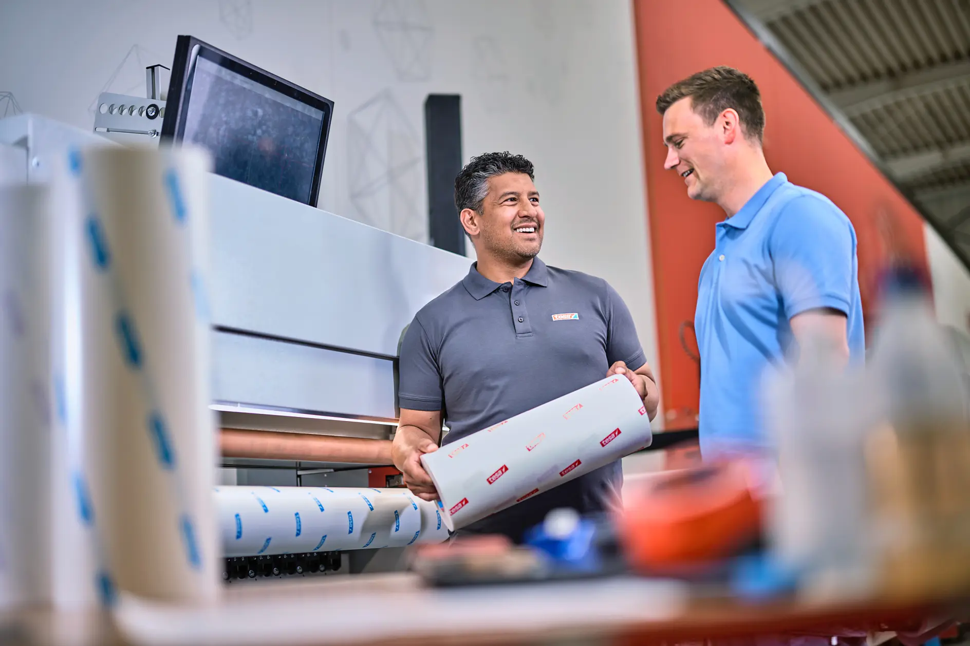 Two men are standing in a printing workshop. One man holds a roll of printed material secured with tesa tape, while the other looks on. A large printer and computer screen are in the background. Both men are dressed in casual work attire. (This text has been generated by AI)