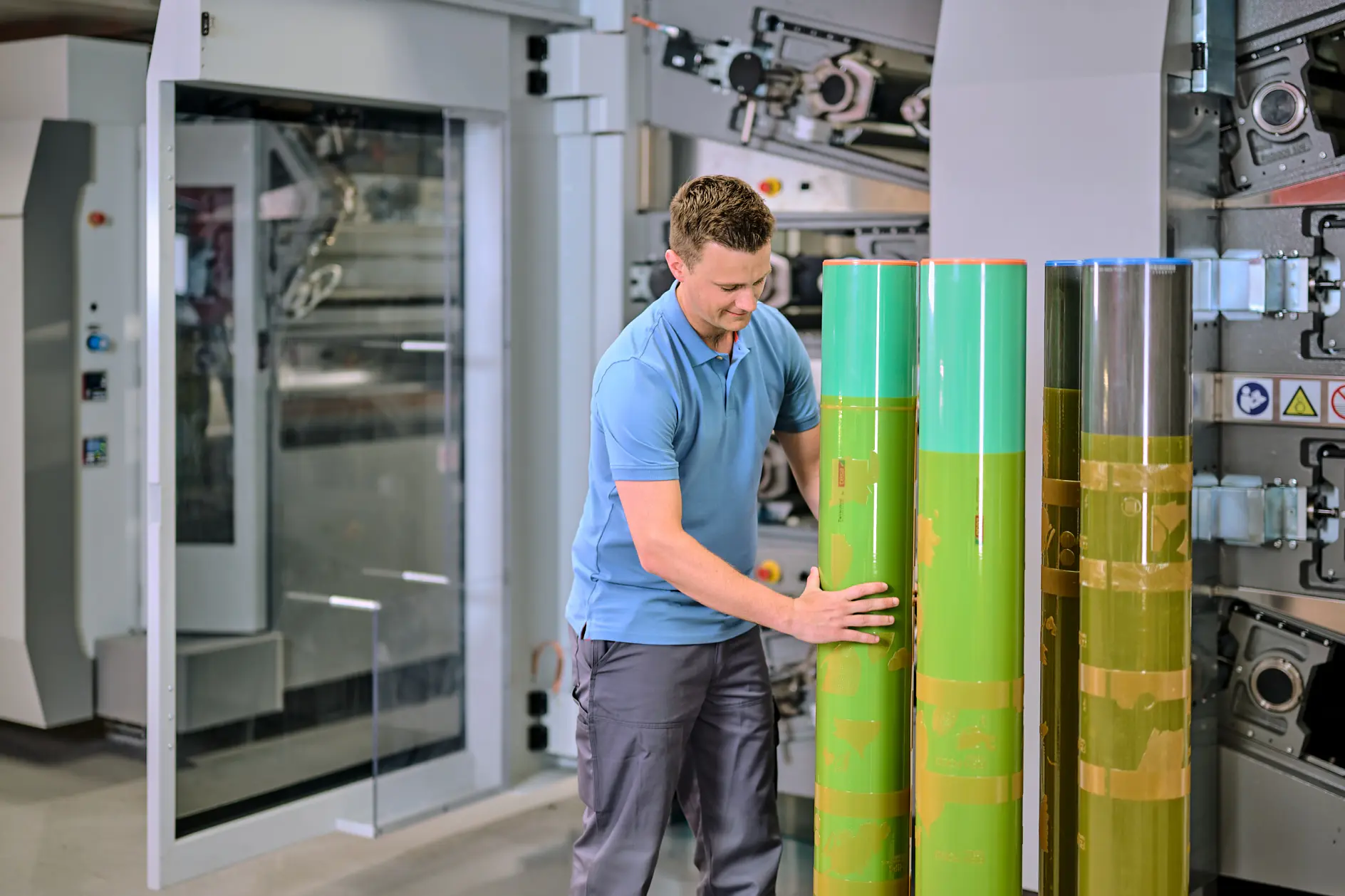 A man in a blue polo shirt and gray pants is inspecting large green cylinders in an industrial setting. He stands in front of a machine with gears and panels, suggesting a manufacturing or printing environment. (This text has been generated by AI)