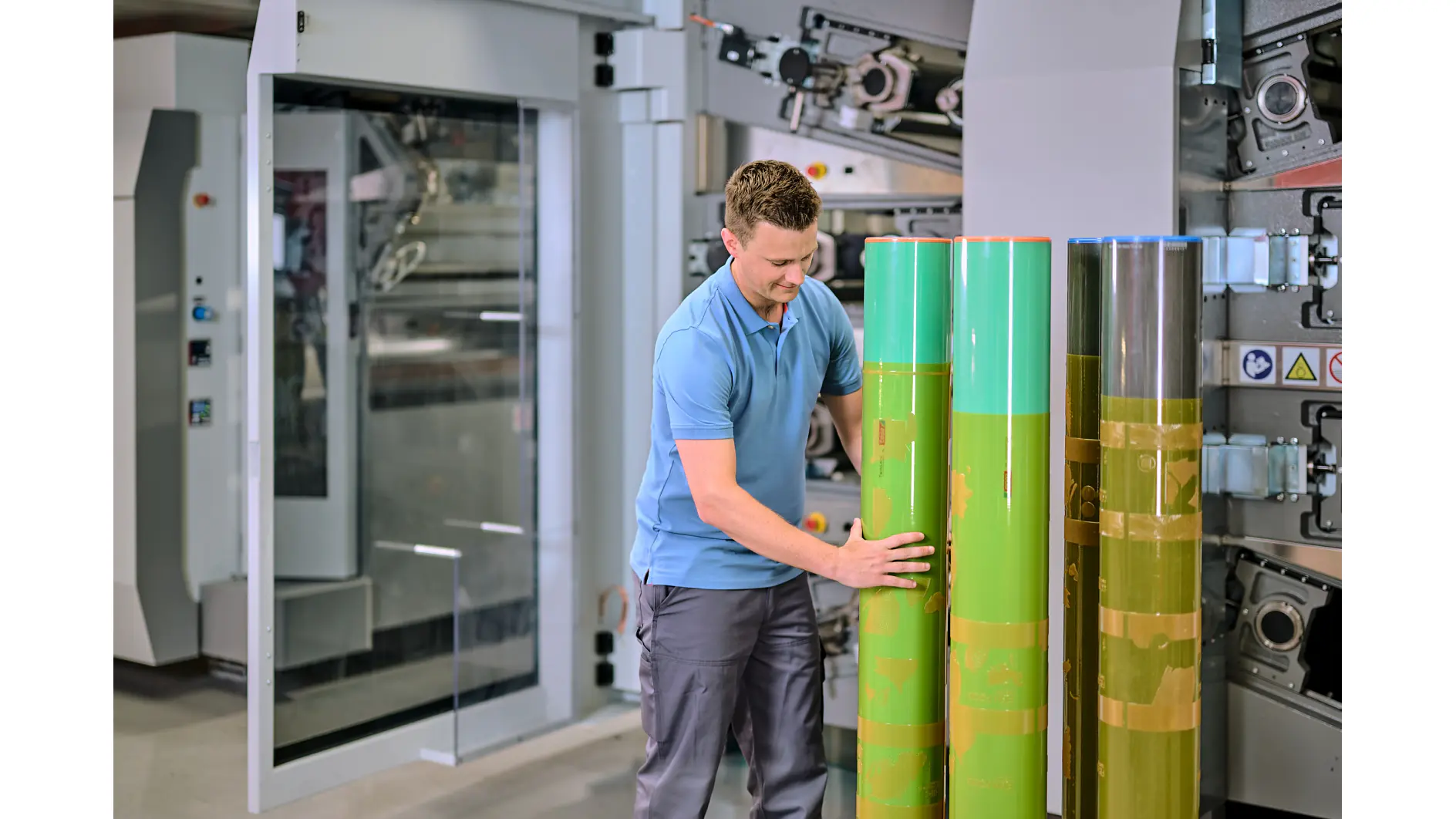 A man in a blue polo shirt and gray pants is inspecting large green cylinders in an industrial setting. He stands in front of a machine with gears and panels, suggesting a manufacturing or printing environment. (This text has been generated by AI)