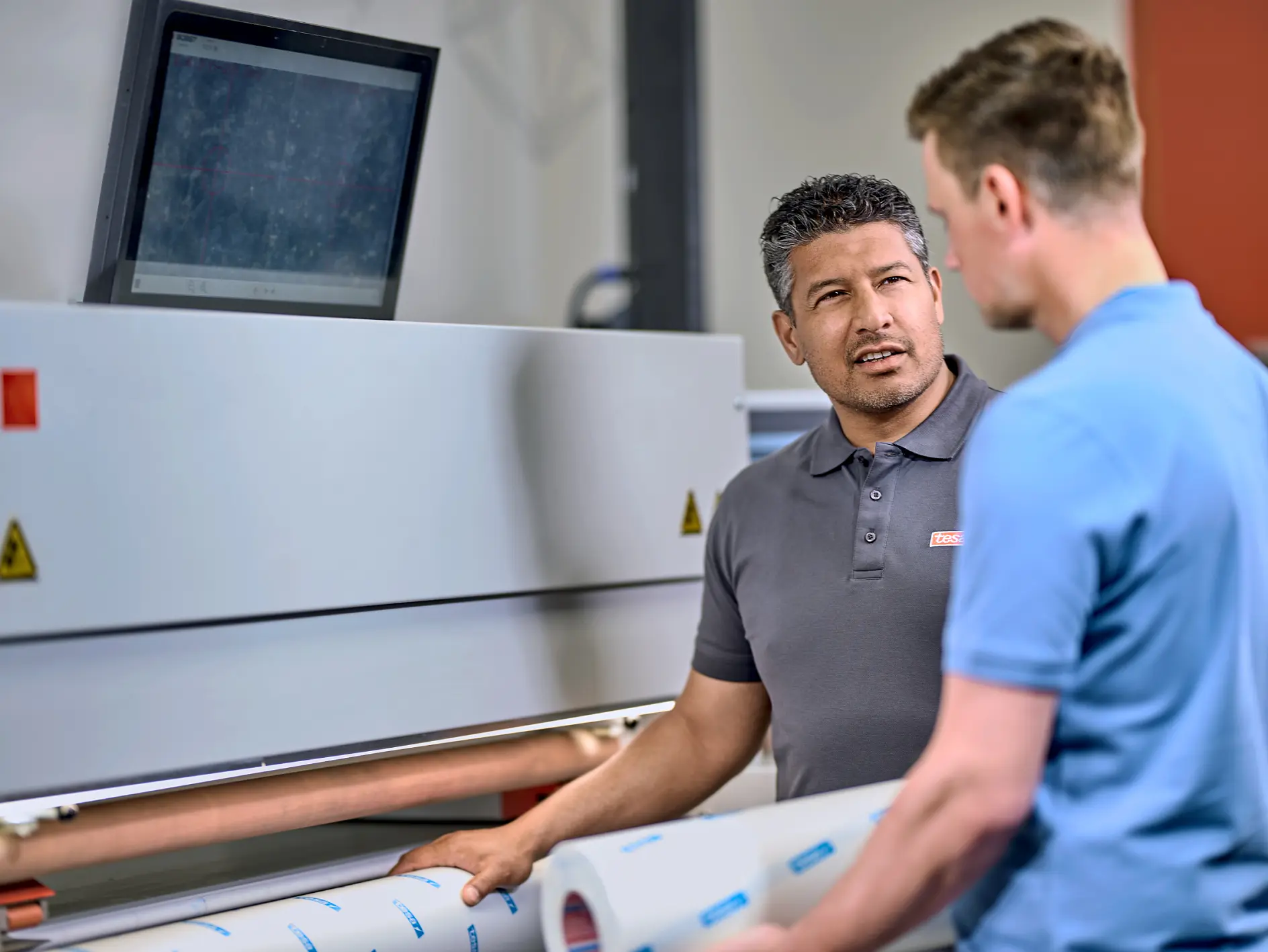 Two men are standing in front of a large industrial machine, discussing something. One man is holding a roll of tesa tape. A computer monitor is mounted on top of the machine. They are in a well-lit workspace. (This text has been generated by AI)