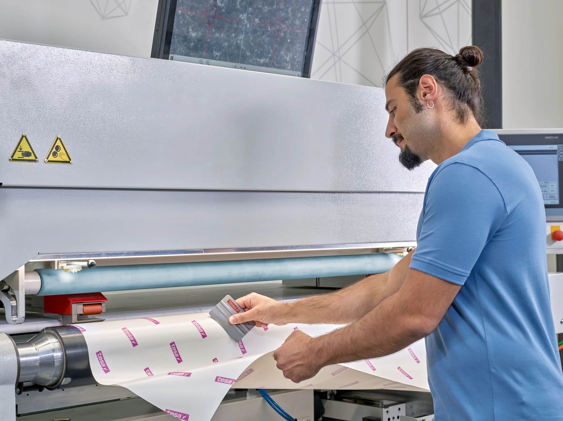 A man in a blue shirt operates a machine, adjusting a piece of paper or material with printed text. A monitor is mounted above the machine, displaying a detailed screen. Control panels are visible, with geometric patterns on the wall. In his hand, he carefully applies tesa tape to secure the material in place, ensuring precision and stability throughout the process. (This text has been generated by AI)