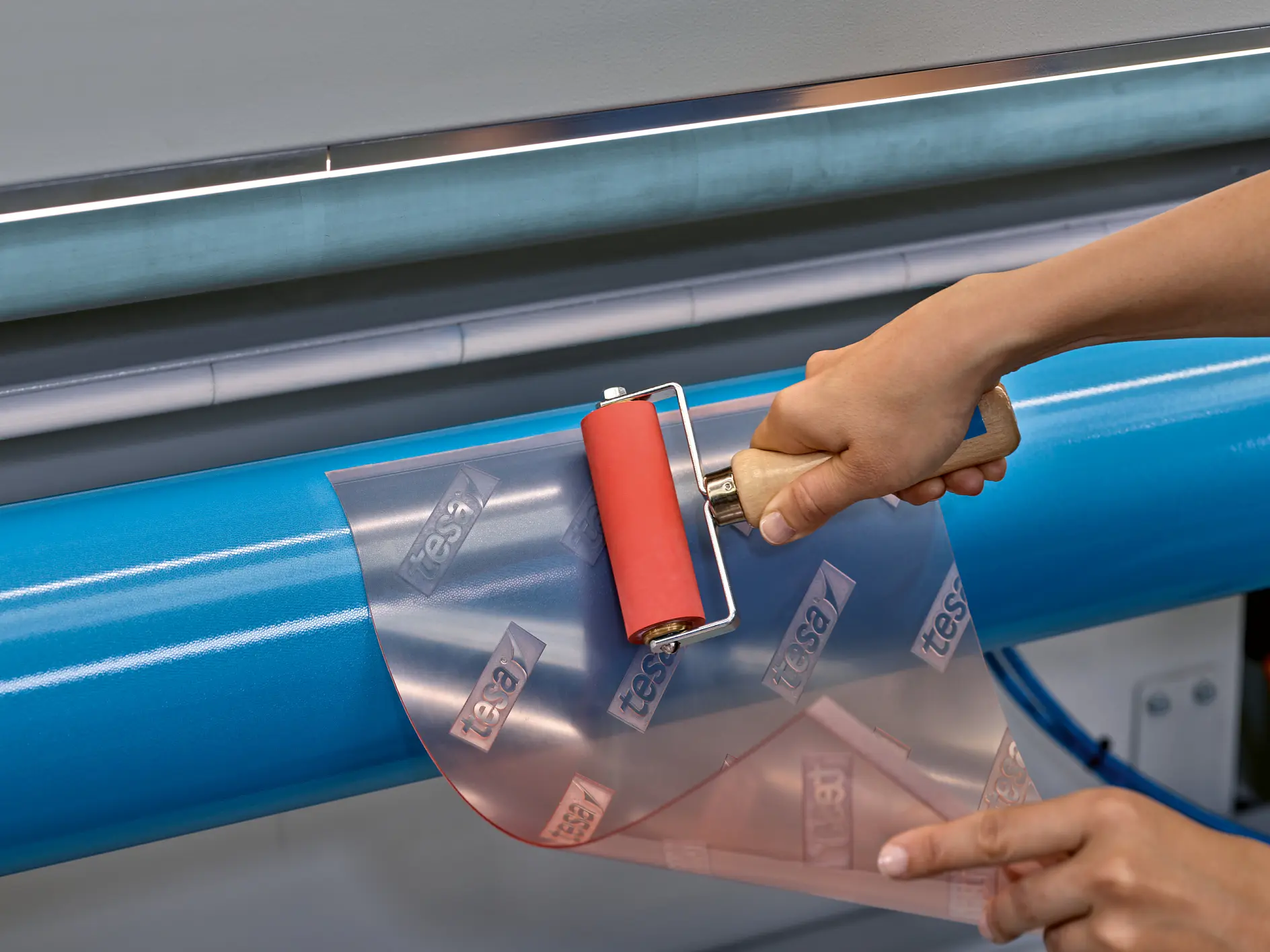 A person uses a red roller to apply clear adhesive film labeled tesa tape onto a blue cylindrical surface. The image focuses on the hands, roller, and film. (This text has been generated by AI)