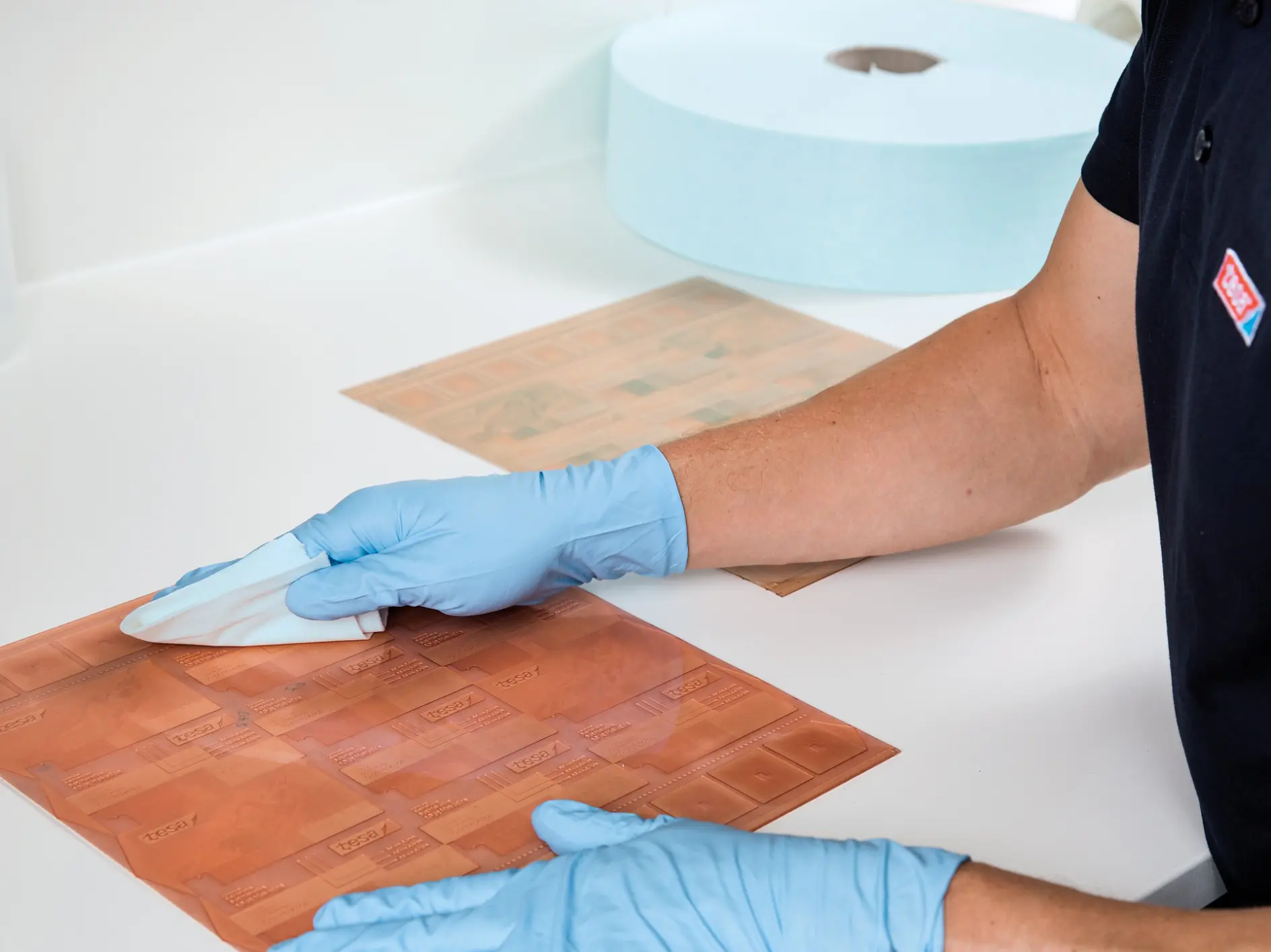 A person wearing blue gloves cleans a rectangular copper plate on a white countertop using a cloth. A bottle of liquid and a large roll of blue tesa tape are visible in the background.
