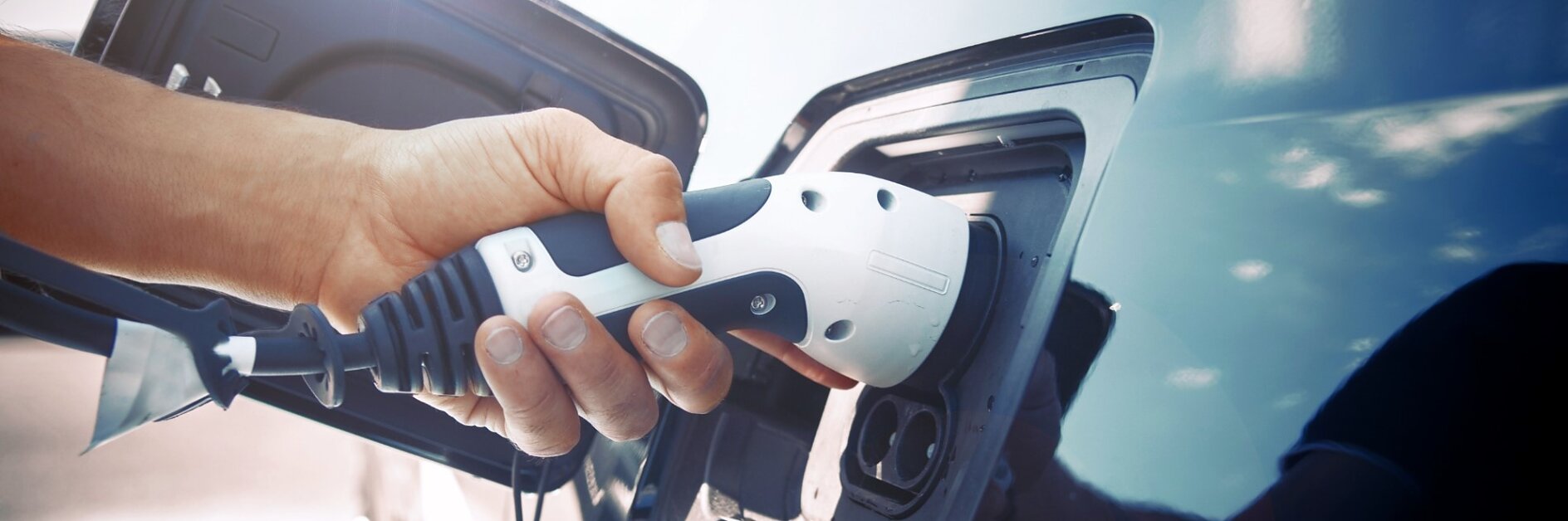 A close-up shows a person plugging a charging cable into an electric vehicle. The hand holds the white connector aligned with the cars charging port, while tesa tape can be seen securing nearby cables. The cars sleek blue exterior is visible in the background.