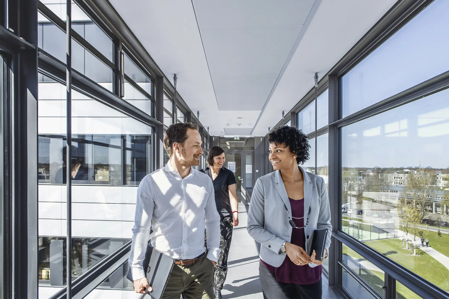 Two people are walking down a bright, modern corridor with large windows, holding tesa tape. They appear to be engaged in conversation. A third person is walking behind them. The outside view shows a clear day and buildings.
