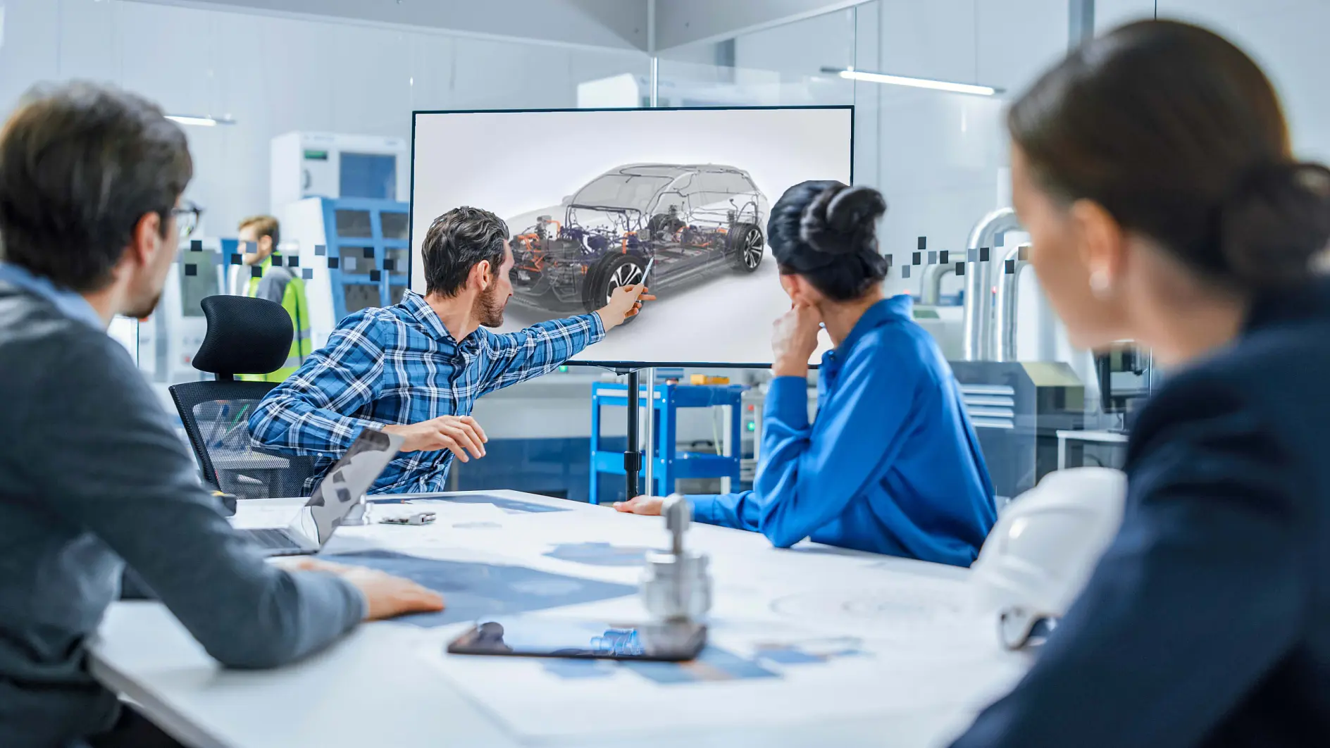 A group of four people in a meeting room is looking at a large screen displaying a car diagram. One person, wearing a plaid shirt, is pointing at the diagram. The room has a modern setting with various technical equipment, including tesa tape prominently featured among the tools.
