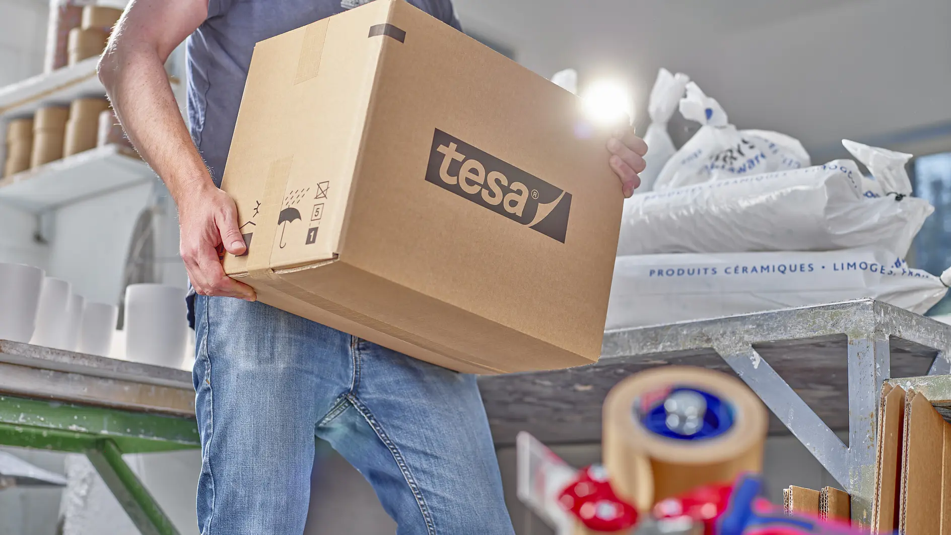 A person holds a large cardboard box labeled tesa in a workspace. Various packing materials are scattered around, including tesa tape dispensers and white sacks. The person is wearing casual clothing, with jeans and a dark top. (This text has been generated by AI)
