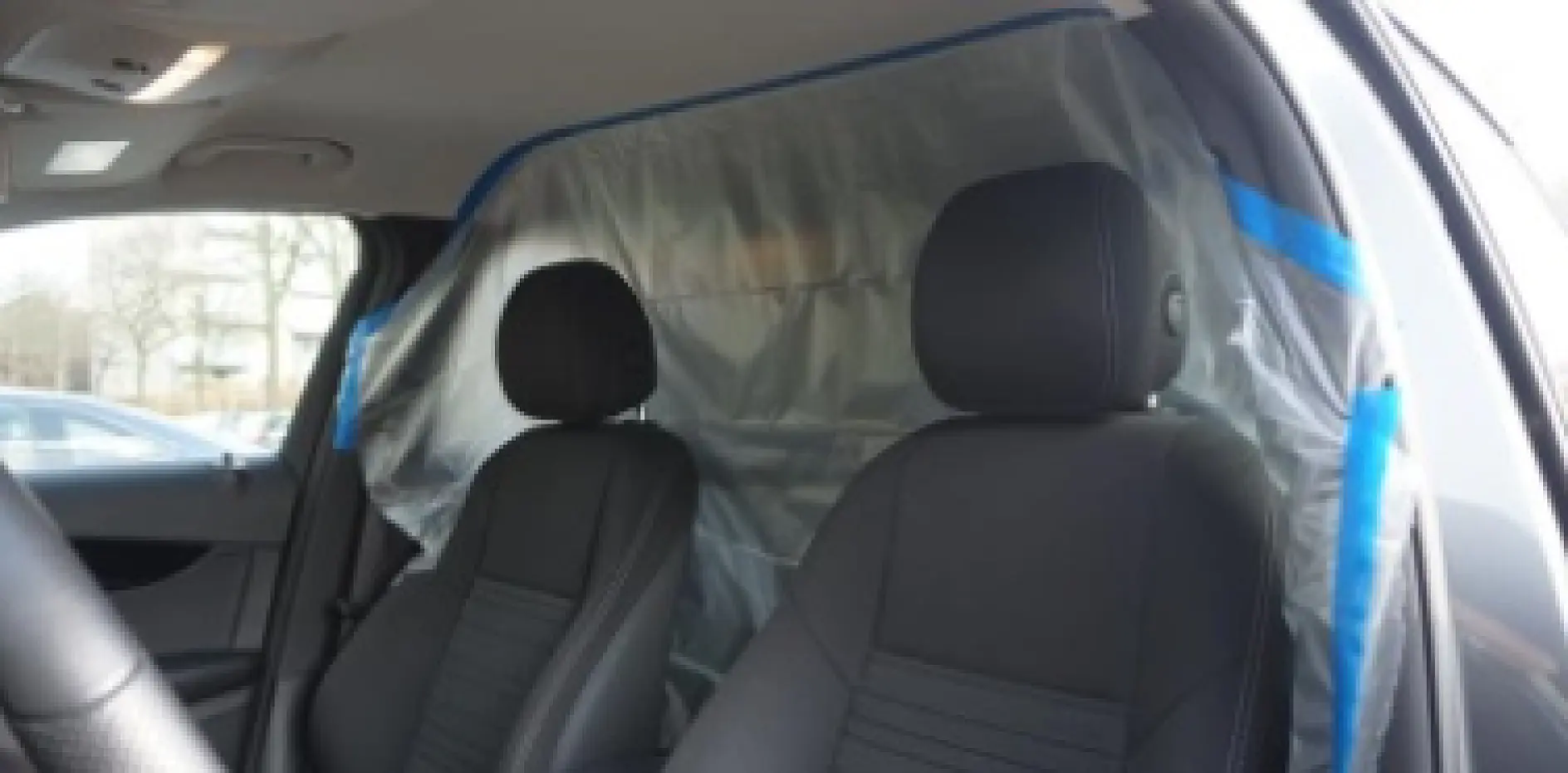 Interior of a car with two front seats covered in black upholstery. A clear plastic sheet is taped with blue tesa tape to create a barrier between the front and back seats. Trees and other cars are visible outside the windows. (This text has been generated by AI)