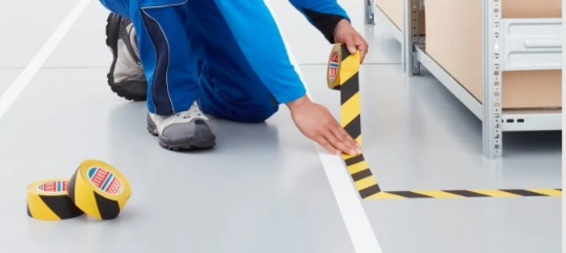 A person in blue overalls is applying black and yellow caution tesa tape to a light gray floor near shelving units. Two additional rolls of tesa tape are on the floor nearby. (This text has been generated by AI)
