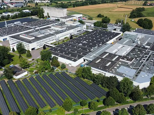 Aerial view of a large industrial complex with multiple white-roofed buildings surrounded by trees, fields, and rows of tesa tape solar panels installed on the ground in the foreground. (This text has been generated by AI)