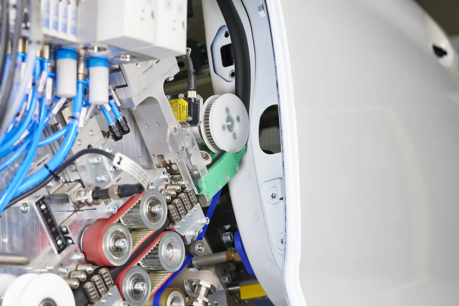 Close-up of an industrial machine with gears, pulleys, belts, blue wires, and tesa tape beside a curved white metal surface.